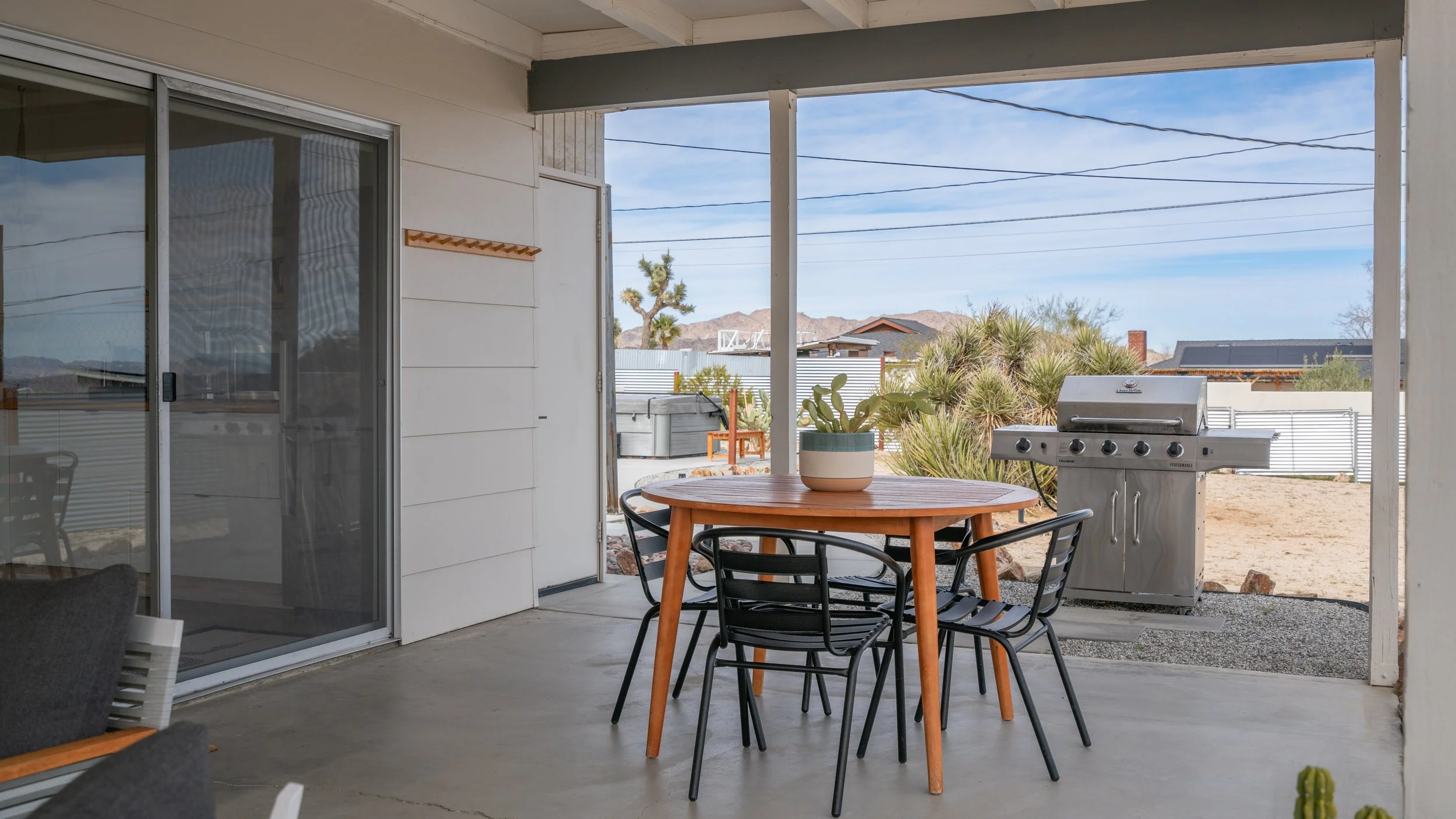 Outdoor patio area with a round wooden table, black chairs, a potted cactus, and a stainless steel barbecue grill. Desert landscaping with cacti and a mountain range in the background.