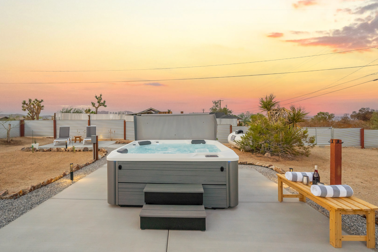 Outdoor hot tub on concrete patio with sunset sky in the background, surrounded by desert landscape, with a wooden bench holding towels and drinks.