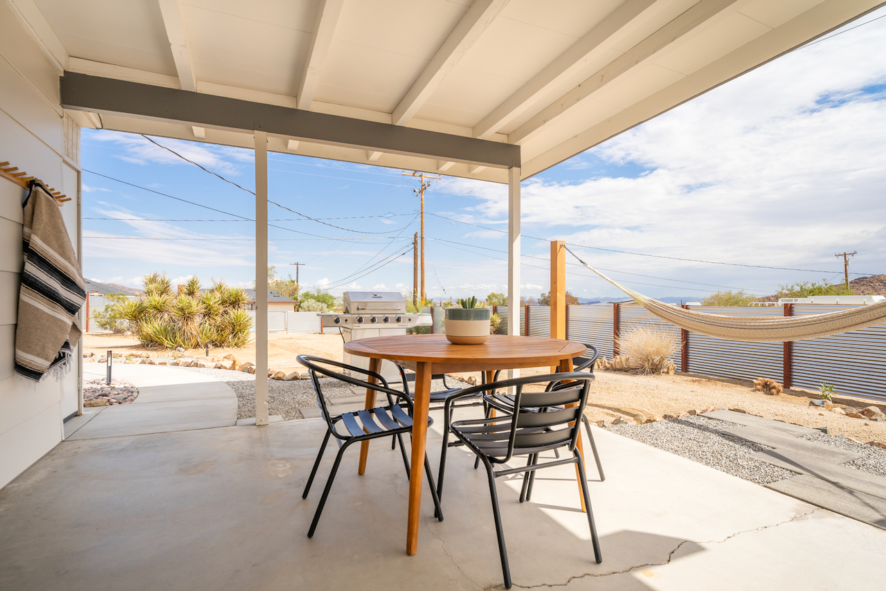 Covered patio with outdoor dining table and chairs, a barbeque grill, a hammock, and desert landscaping with cacti under a partly cloudy sky.