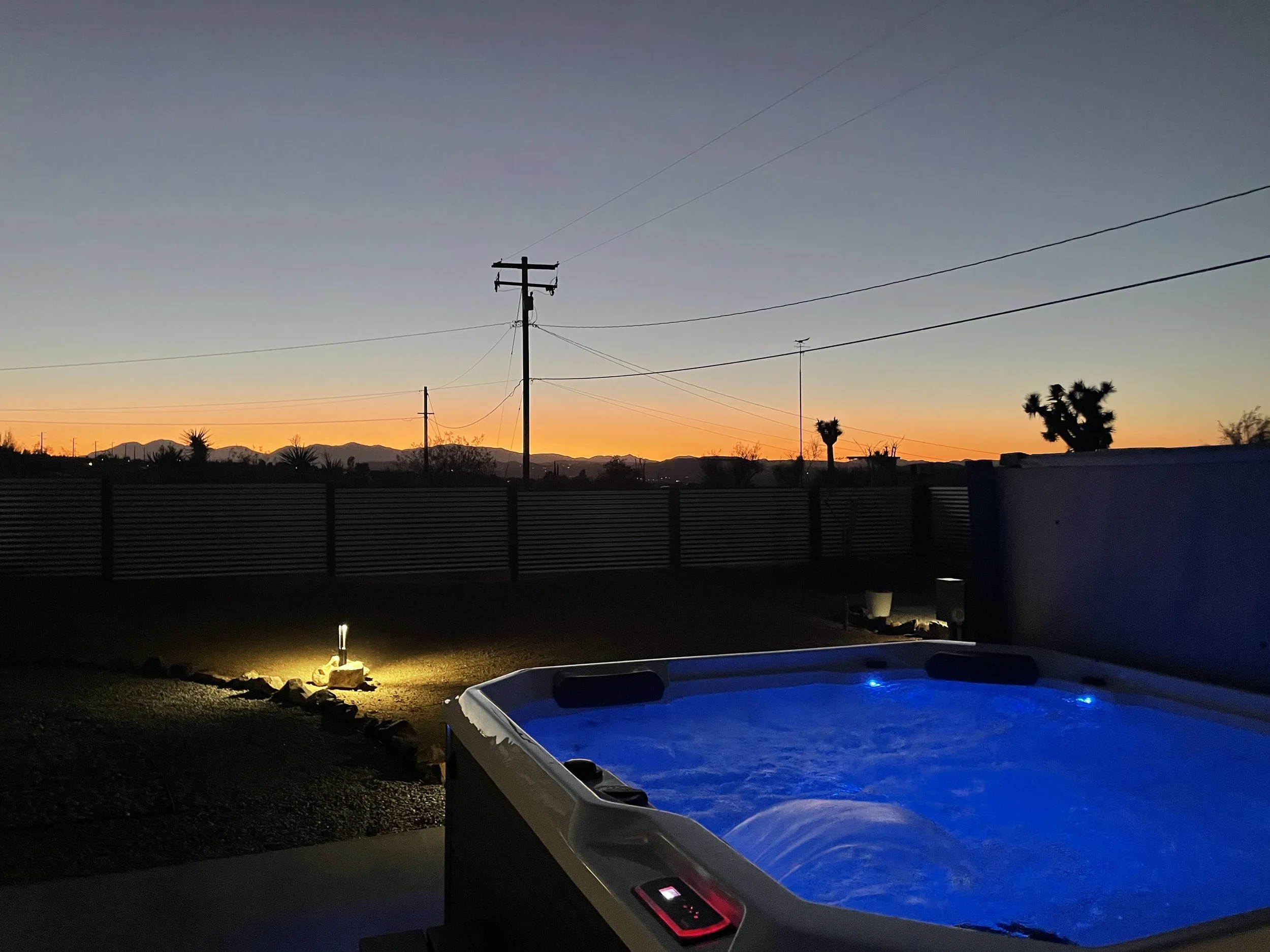 A hot tub with blue lights in a backyard during sunset, with a fence, power lines, and mountains in the background.