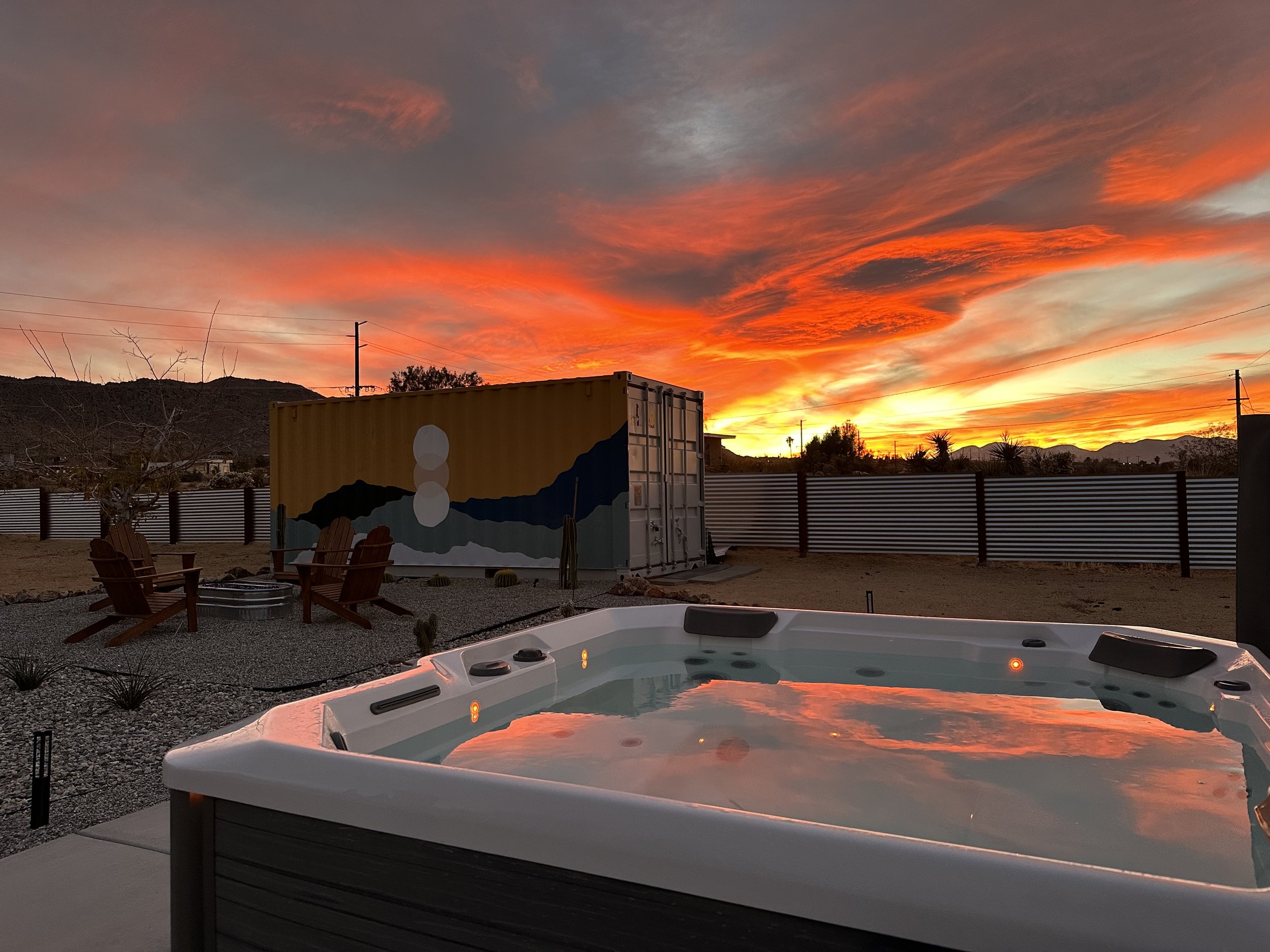 A backyard with a hot tub, four wooden chairs around a fire pit, a large shipping container painted with mountains and a sunset, under a dramatic colorful sunset sky with orange, pink, and purple clouds, and a metal fence in the background.