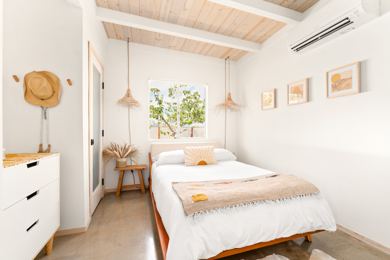 A cozy bedroom with white walls and a wooden ceiling, featuring a bed with white linens and a decorative pillow, two hanging pendant lights, a small wooden side table with a plant, a window showing greenery outside, and framed artwork on the wall.