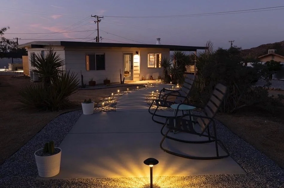 A small house with string lights along the walkway, outdoor chairs on a concrete patio, potted plants, desert plants nearby, and a lit landscape light in the evening.