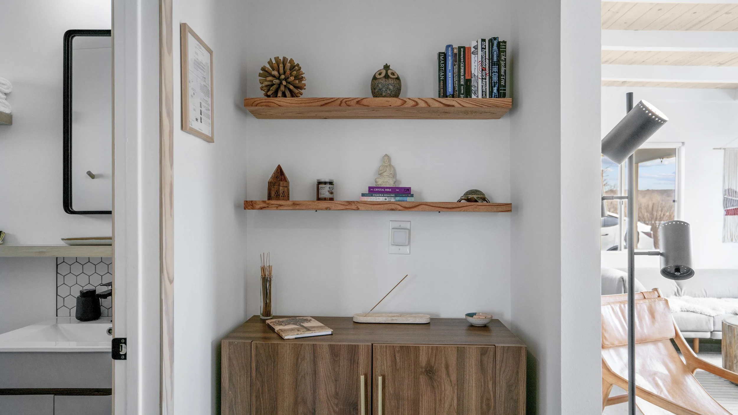 Interior of a room with two wooden shelves holding books and decorative items, a wooden cabinet with a book, reed diffuser, dish, and incense stick on top, and a houseplant in a vase beside it, with part of a bathroom visible on the left.