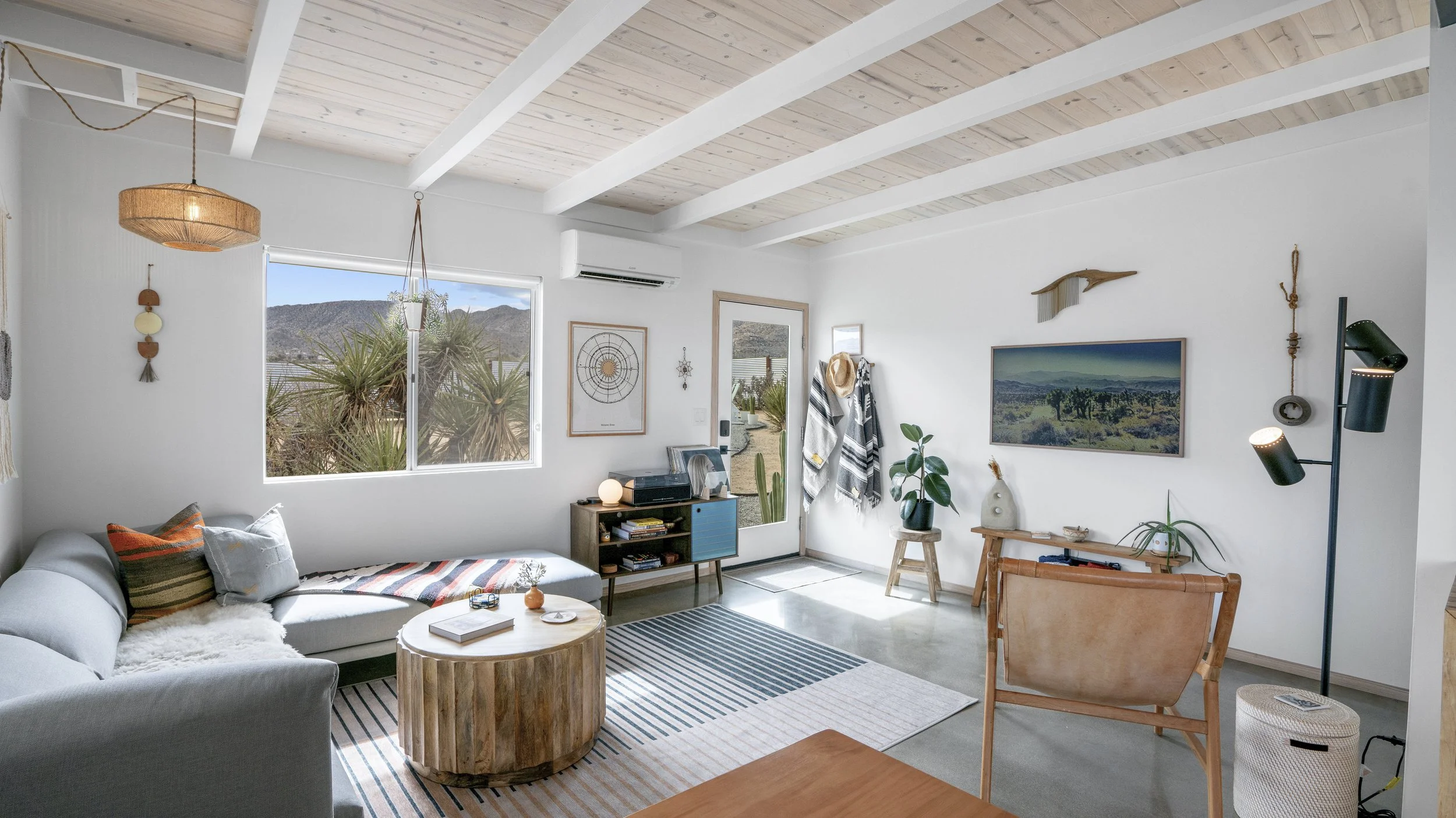 Bright living room with white walls, wooden ceiling beams, a large window showing desert landscape, a gray sectional sofa with pillows, a wooden coffee table, a small blue entertainment console, a wall-mounted flat-screen TV, indoor plants, and decor