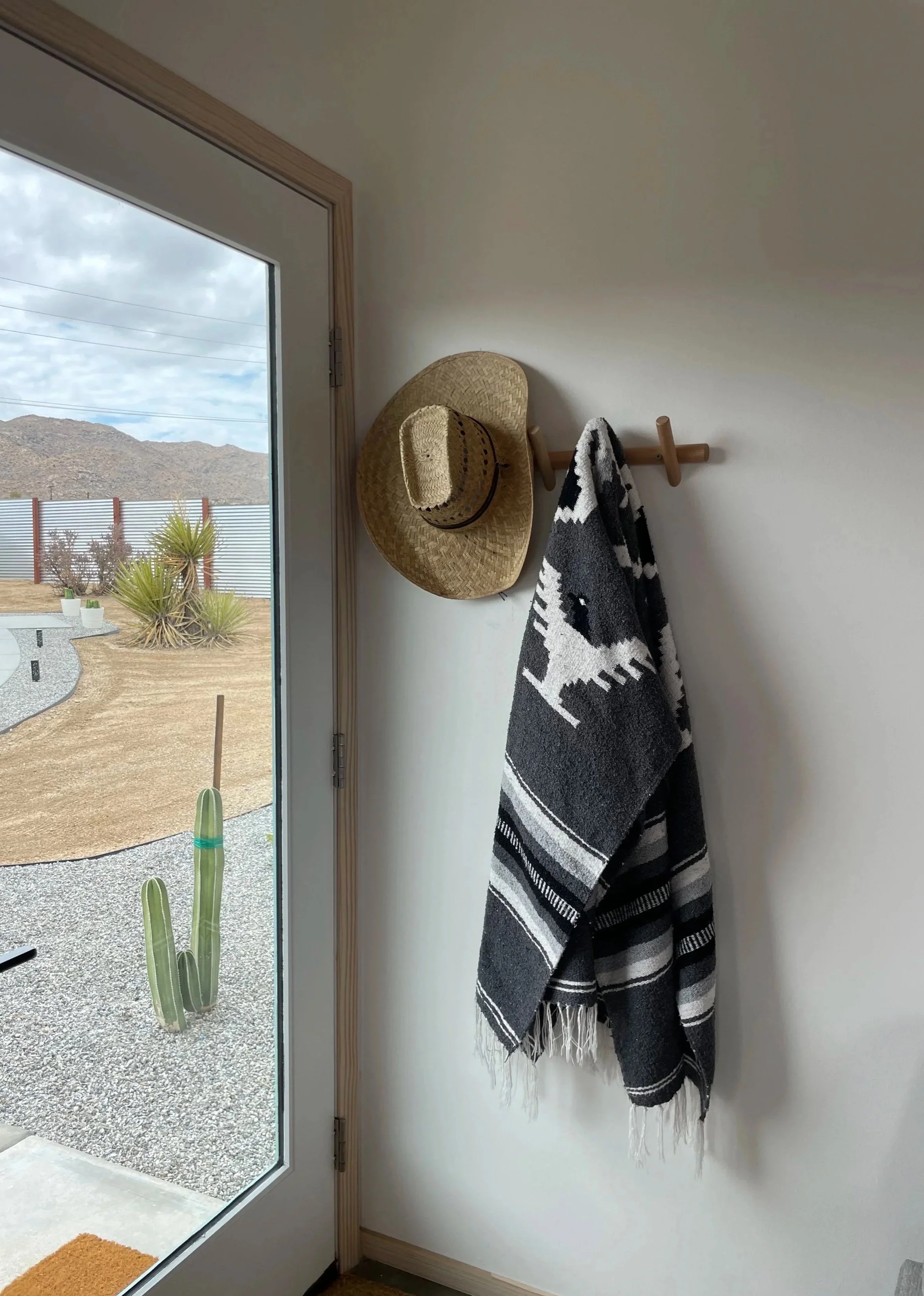 Interior corner with coat rack holding a straw hat and a black and white striped blanket near a glass door, with desert landscape and cactus outside visible through the door.