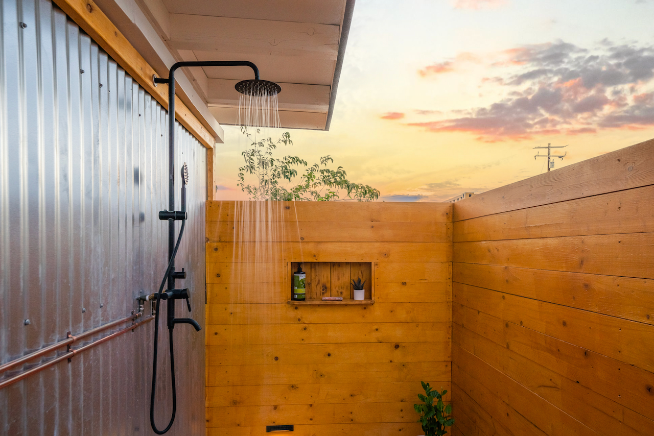 Outdoor shower with rain shower head mounted on a metal pipe, wooden privacy wall, sunset sky with clouds, small shelf with soap, plant, and bottle, and additional plants at the bottom.