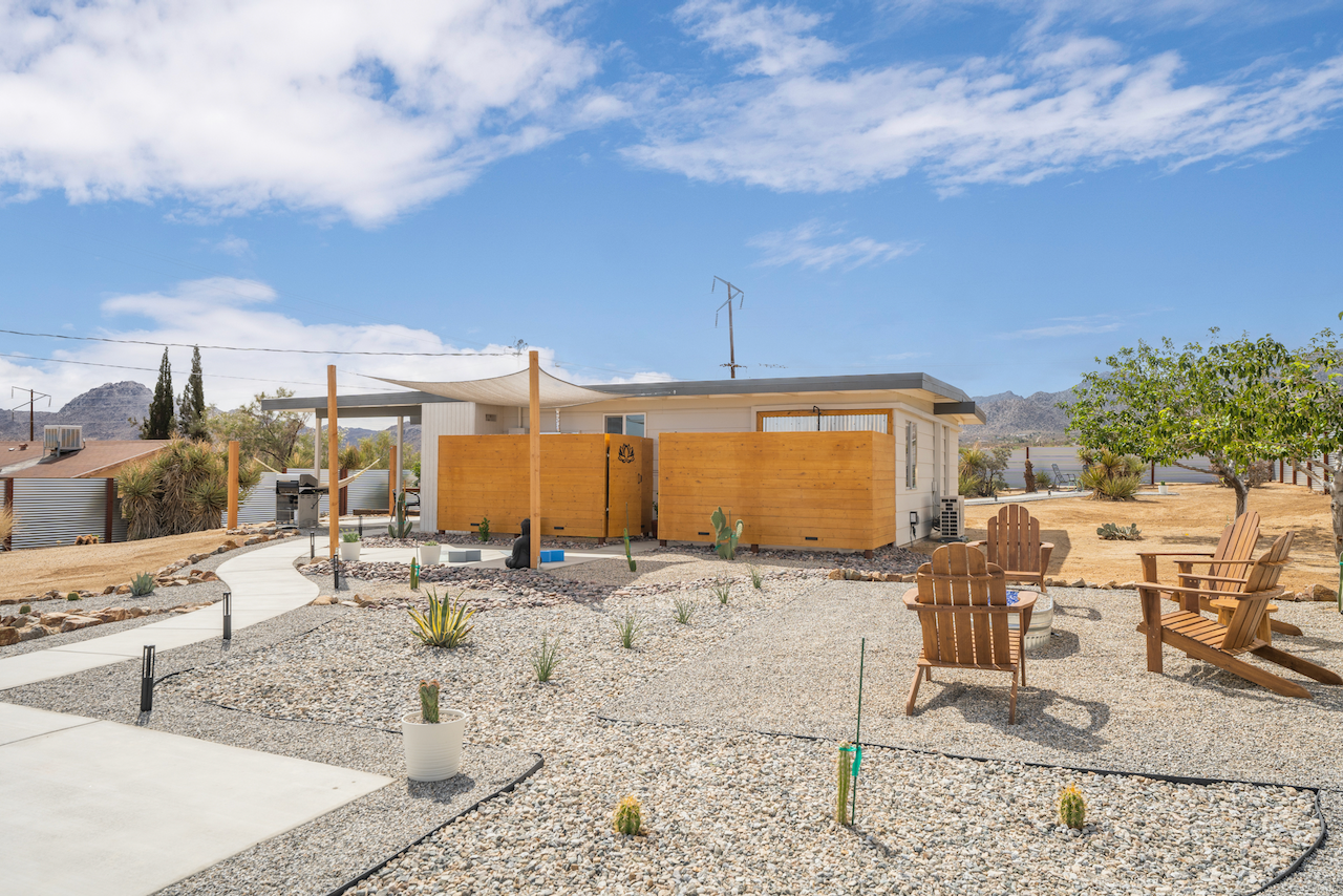 Backyard with gravel ground, wooden chairs around a small fire pit, cactus plants in pots, a pathway, a small house with outdoor furniture, trees, and mountains in the background on a sunny day