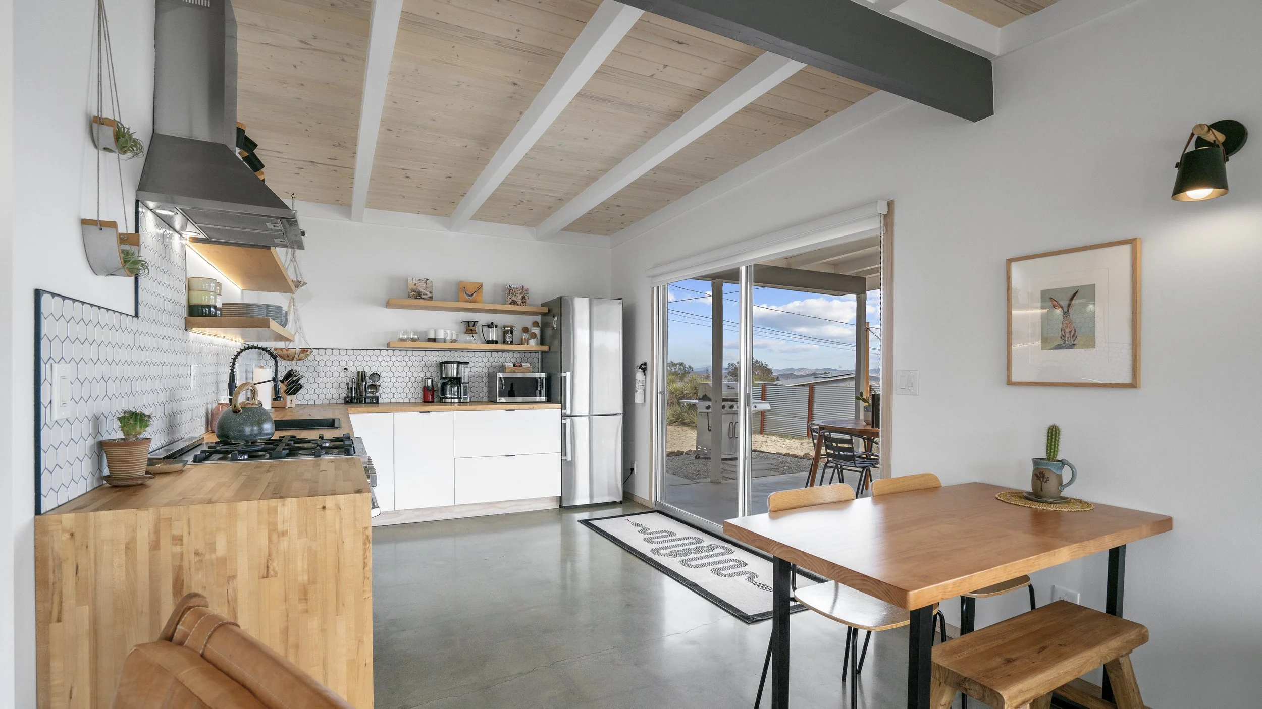 Modern kitchen with white cabinets, open wooden shelves, black hexagon tile backsplash, stainless steel refrigerator, and wooden dining table with bench seating, natural light from sliding glass door, and minimalist decor.