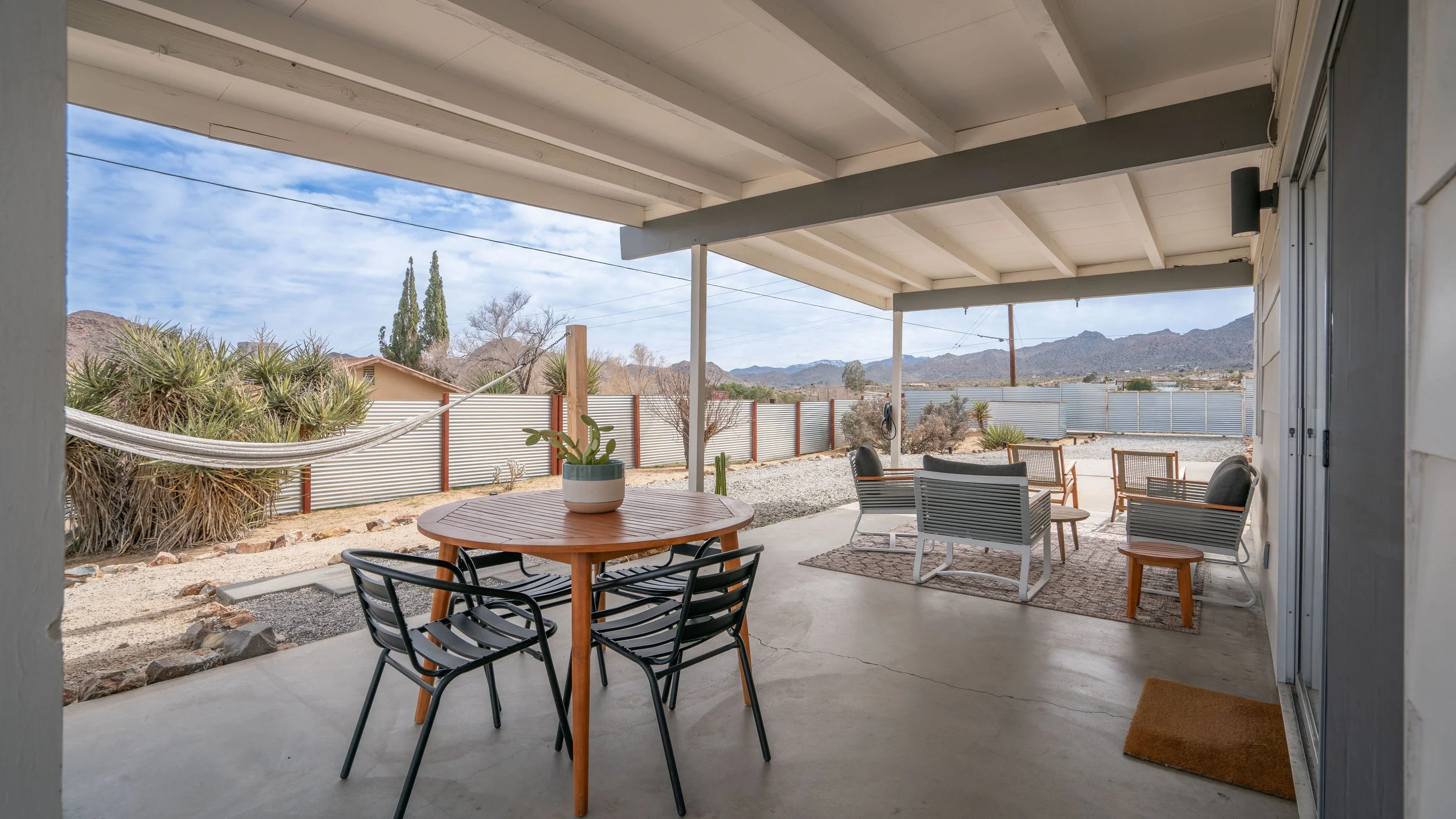 Covered patio with outdoor furniture including a round table with a potted plant, lounge chairs, and mountain views in a desert landscape.