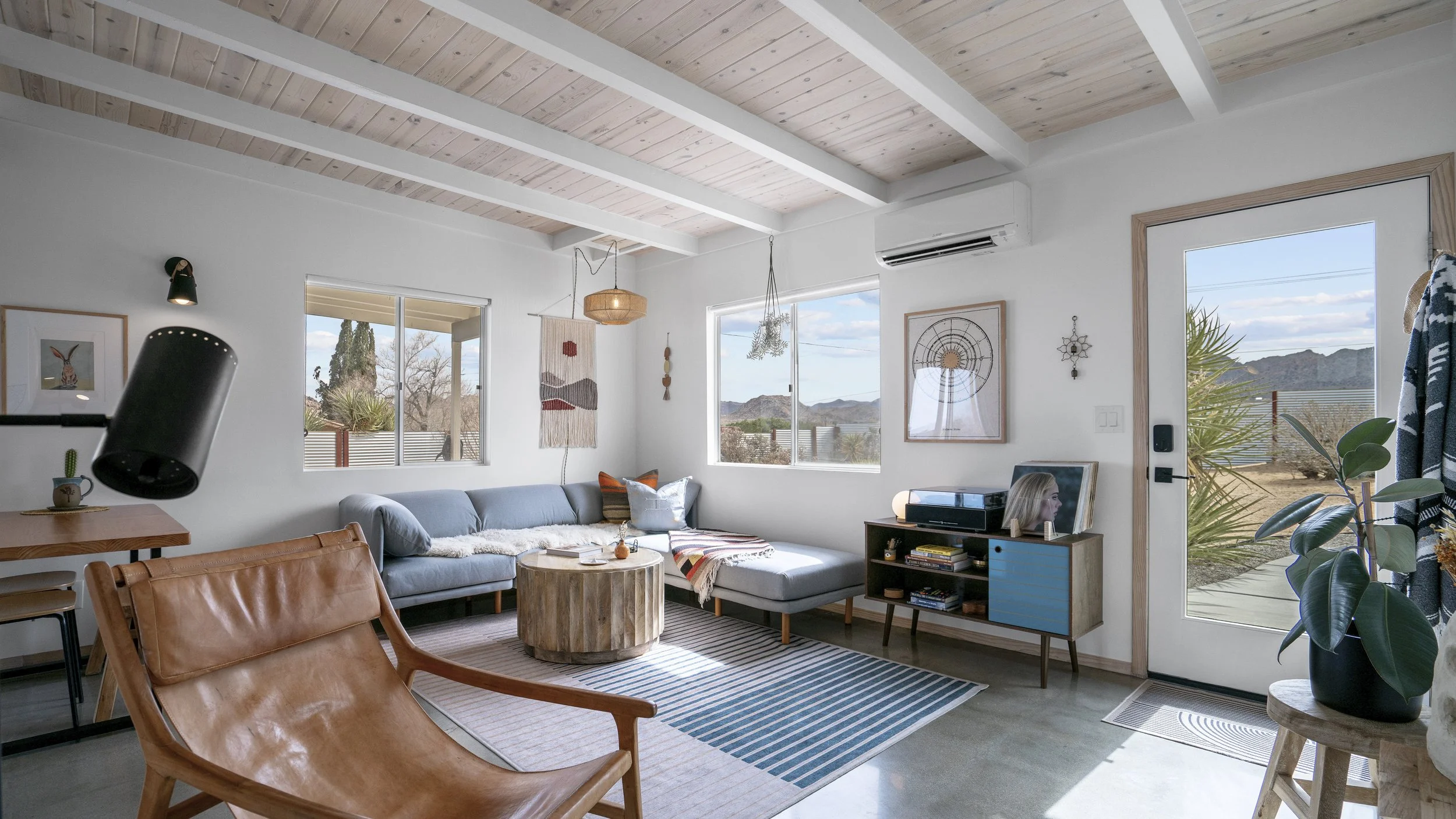 A cozy living room with white walls, large windows showing a desert landscape, and a wooden ceiling. There's a light blue sectional sofa with colorful pillows, a round wooden coffee table, and a leather armchair. Decor includes wall art, plants, and 