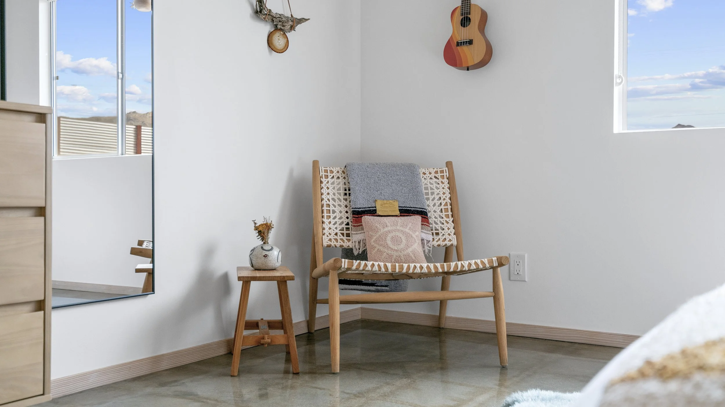 A cozy corner with a wooden chair draped with colorful blankets and pillows, a small wooden side table with a vase of dried flowers, a wall with hanging art, and two windows showing a blue sky with clouds.