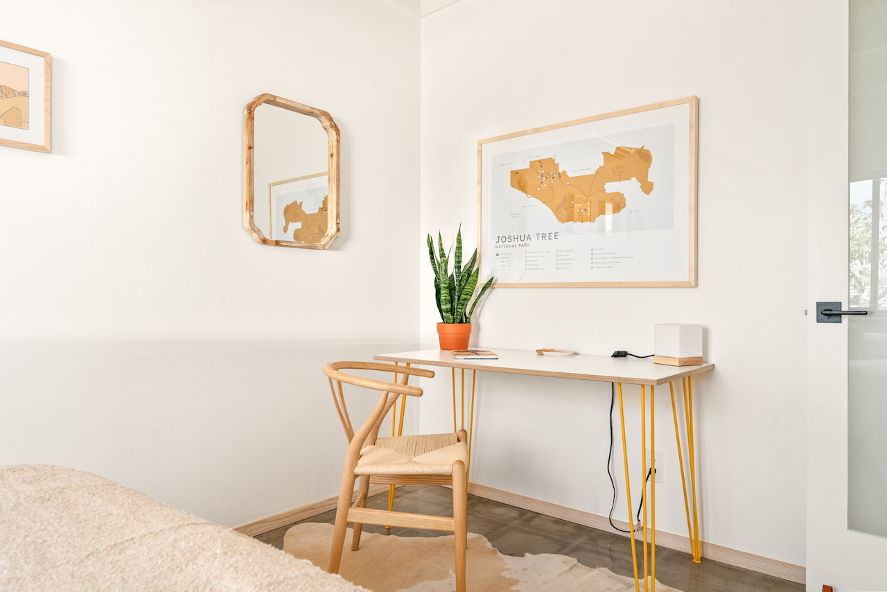 A minimalist home office with a wooden desk and chair, a potted snake plant, artwork on the wall, and a mirror. The desk has a small lamp, a book, and an electronic device. The decor is light and modern with neutral tones.