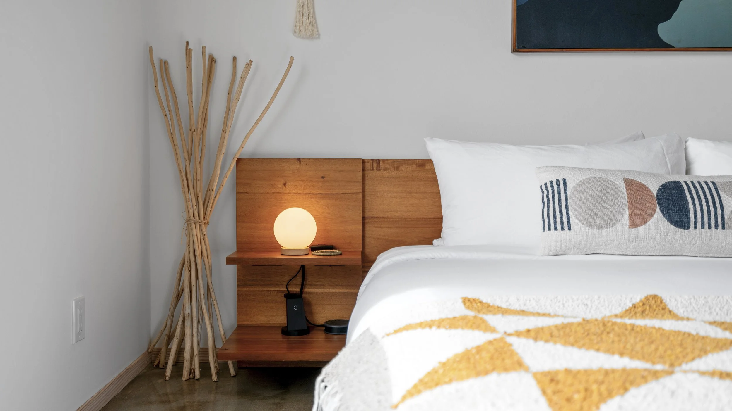 Bedroom corner with wooden headboard, white bedding, geometric decorative pillow, bedside table with glowing lamp and electronic devices, and tall dried twig arrangement in the corner.