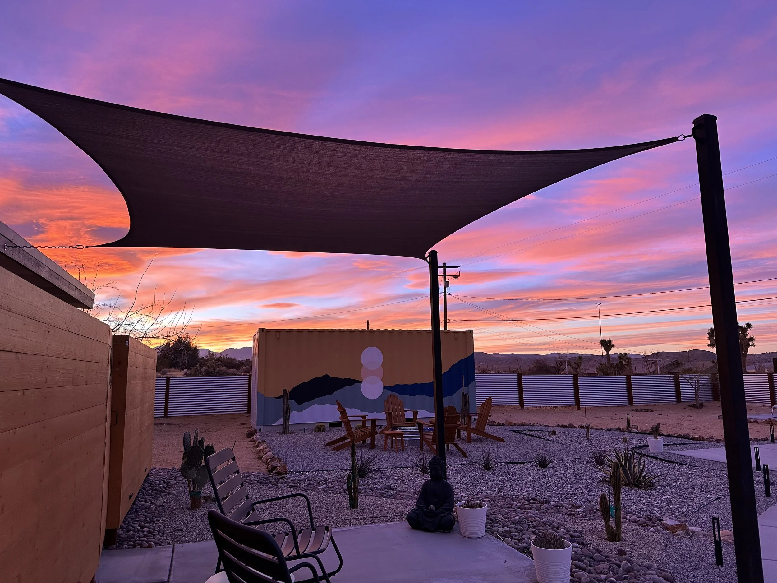 Sunset sky with pink and purple clouds over a backyard patio with seating, cacti, and a painted mural wall, shaded by a large triangular shade sail.