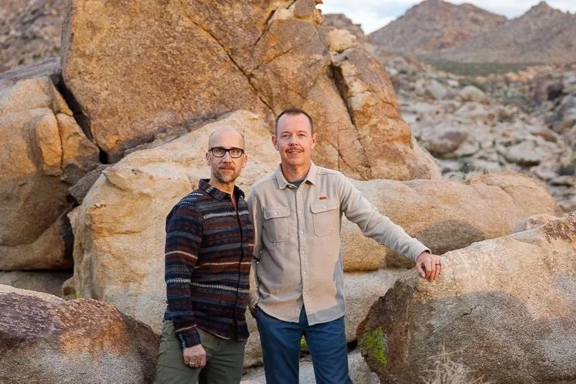 Two men standing between large rocks in a desert landscape with mountains in the background, one wearing a striped sweater and glasses, the other in a light-colored shirt, smiling.