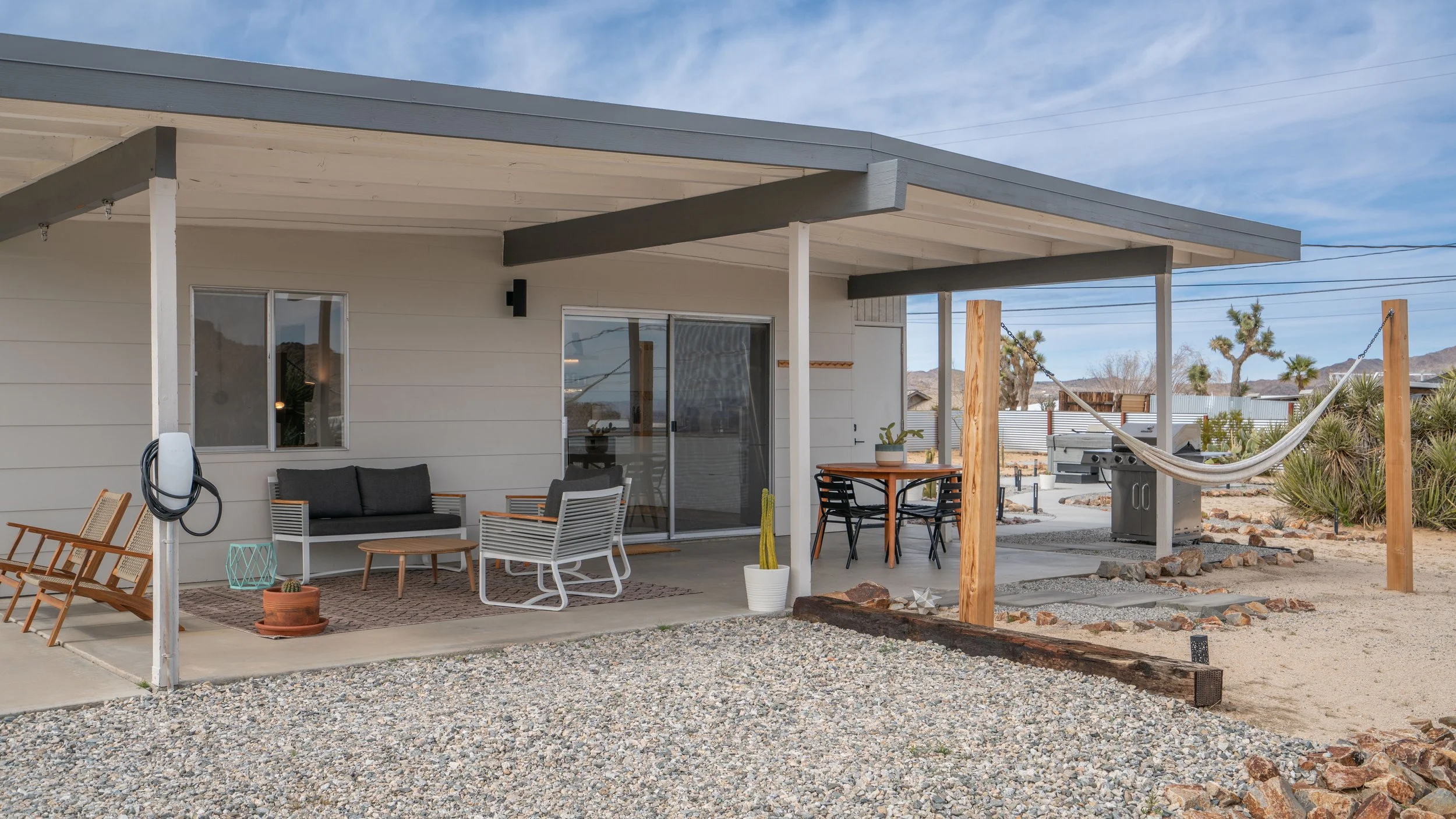 Back porch of a house with outdoor seating, potted plants, a hammock, and a grill in a desert landscape.