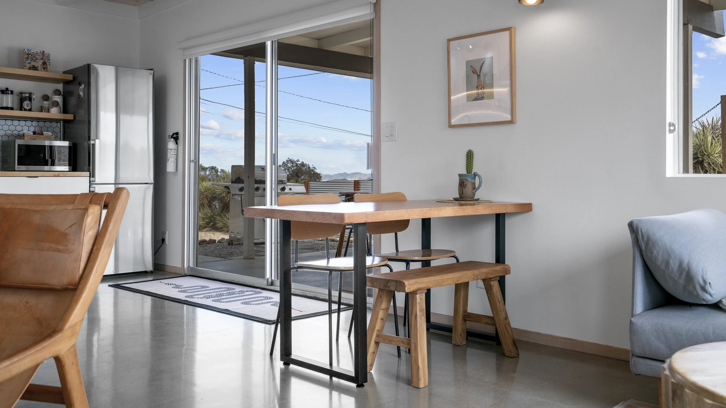 Living room with a wooden dining table, chairs, a gray cushion sofa, a decorated wall with a framed illustration, a potted cactus, and a sliding glass door leading to an outdoor patio with a barbecue grill, overlooking a landscape with trees and moun
