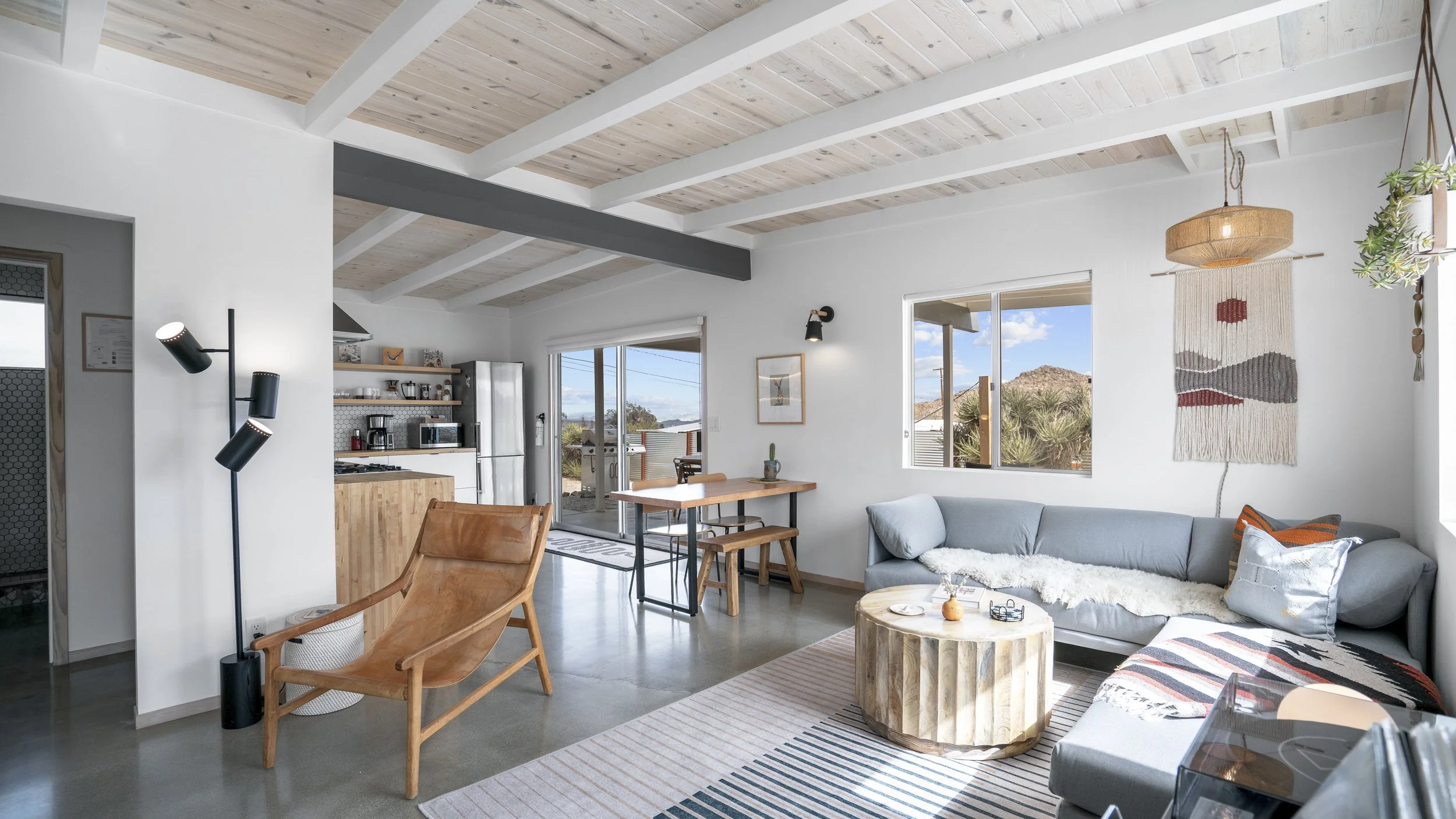 Living room with white walls, a light gray corner sofa with pillows, a round wood coffee table, a striped rug, and a wall hanging. Open kitchen in the background with stainless steel refrigerator and open shelves. Large windows and a sliding door lea