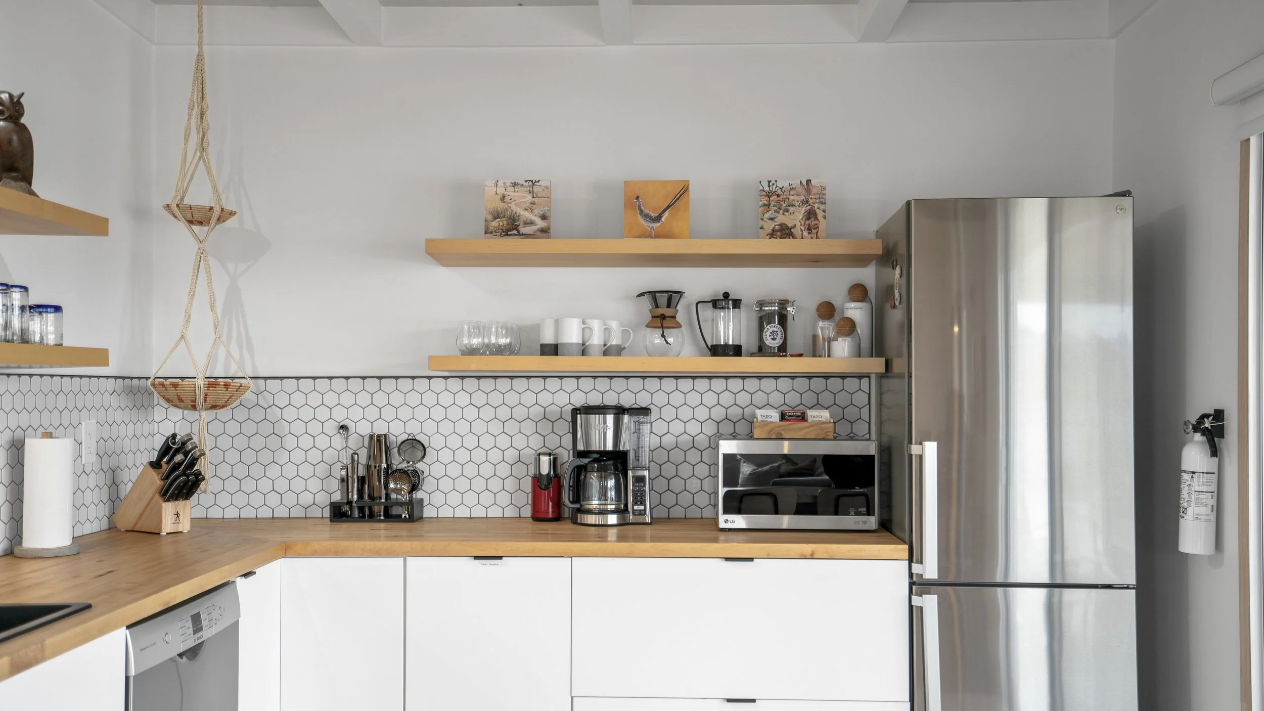 Modern kitchen with white cabinets, wooden countertop, and stainless steel refrigerator. Open wooden shelves hold art prints and kitchenware. The backsplash features white hexagonal tiles. Various appliances and utensils are arranged on the counter.