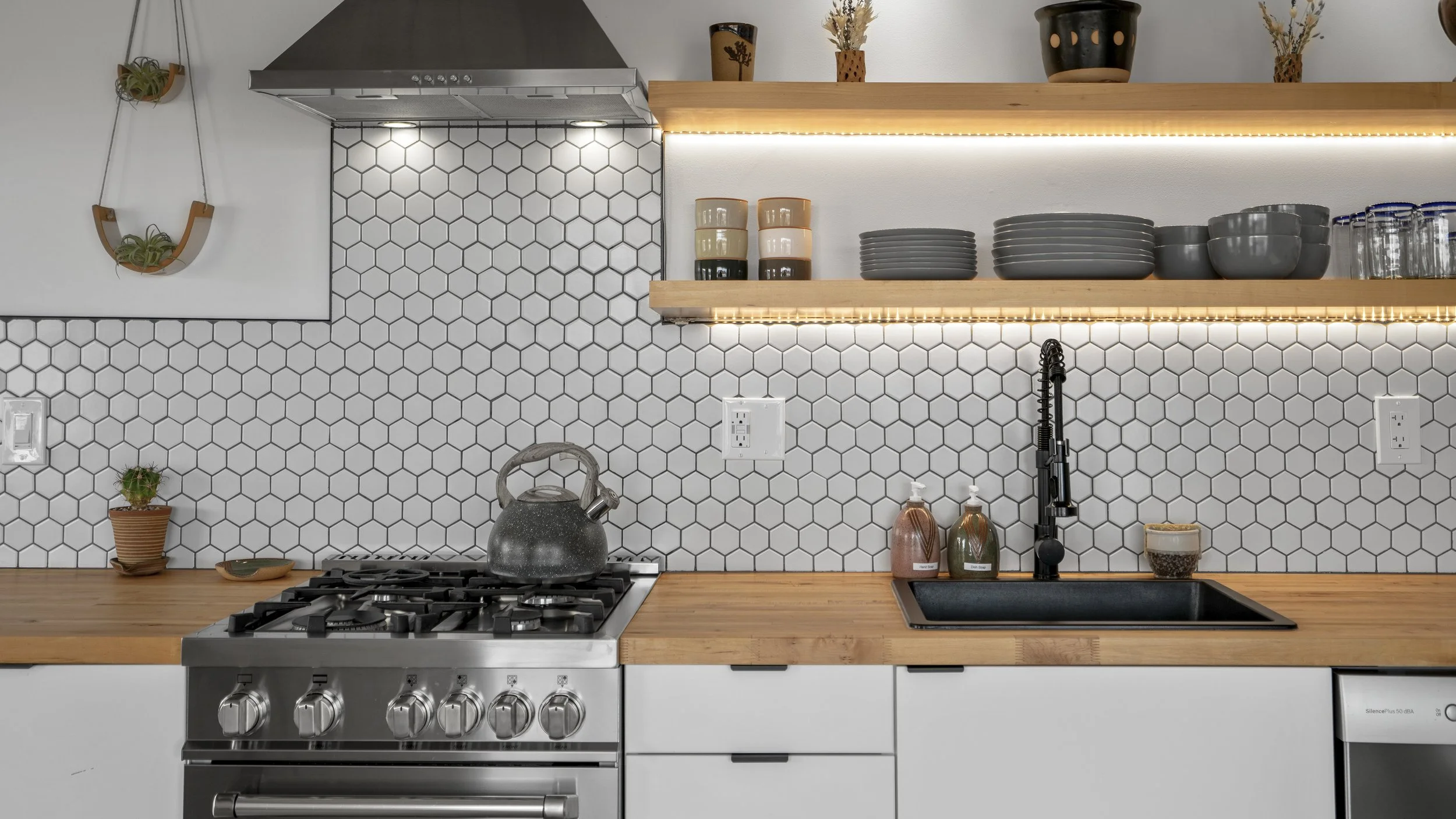Modern kitchen with white hexagonal tile backsplash, wooden countertop, open shelves with dishes and decor, black sink, and a stainless steel stove with a tea kettle.
