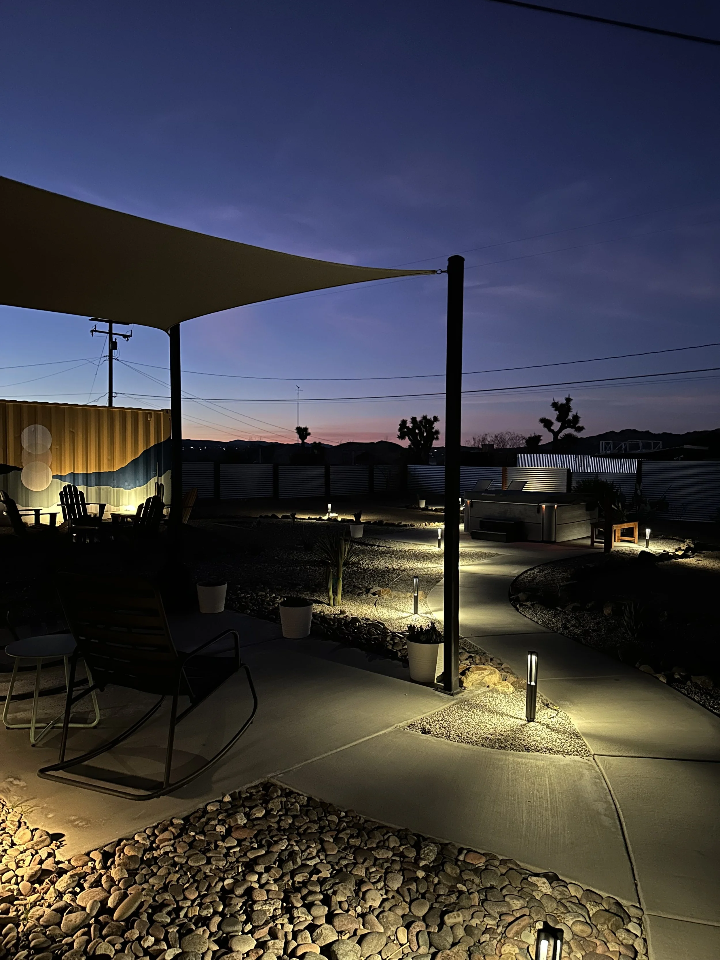 A backyard patio at dusk with outdoor lighting, modern chairs, potted plants, a hot tub, and a sail shade overhead. Mountains and a few trees silhouette in the background.