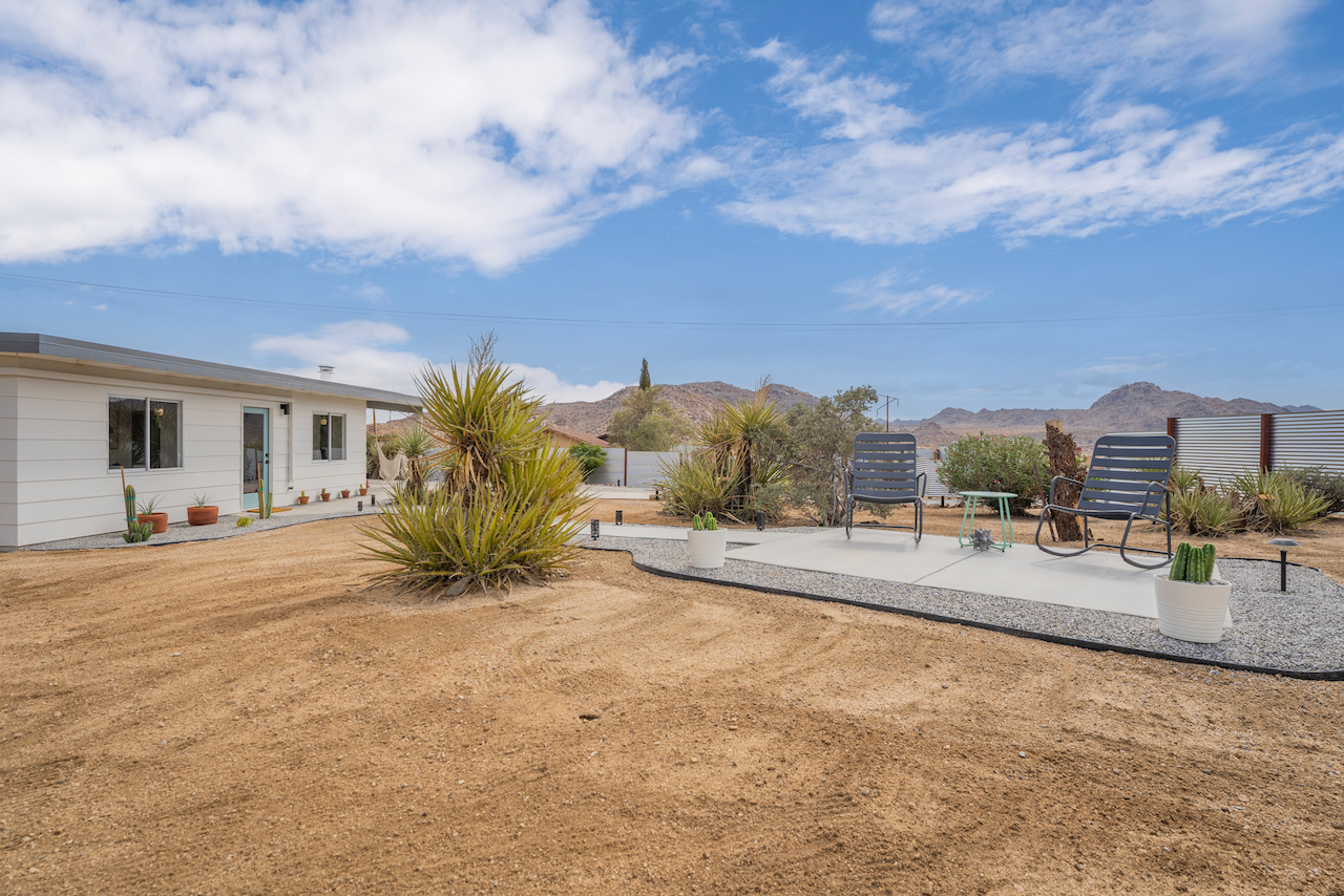 Desert backyard with a house on the left, a paved sitting area with metal chairs and a small table, potted cacti, and desert vegetation under a partly cloudy sky, with mountains in the background.