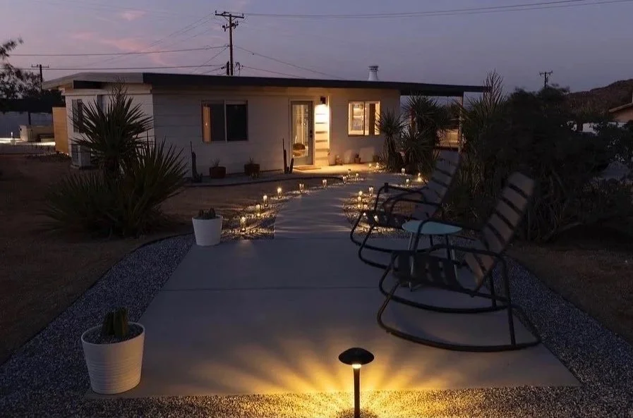 A cozy backyard patio at dusk with a concrete pathway lined with small candles, leading to a white house with warm lighting. There are potted plants and two lounge chairs on the right side.