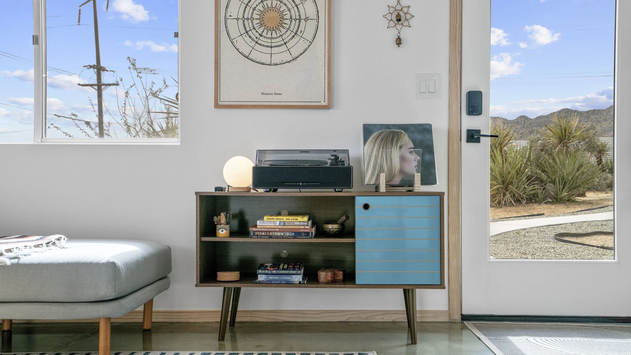 Interior of a modern home featuring a wooden sideboard with books, a turntable, and decor placed on top. A lamp emits warm light, and a mirror reflects a profile of a woman. There are large windows and a glass door showing a desert landscape with cac