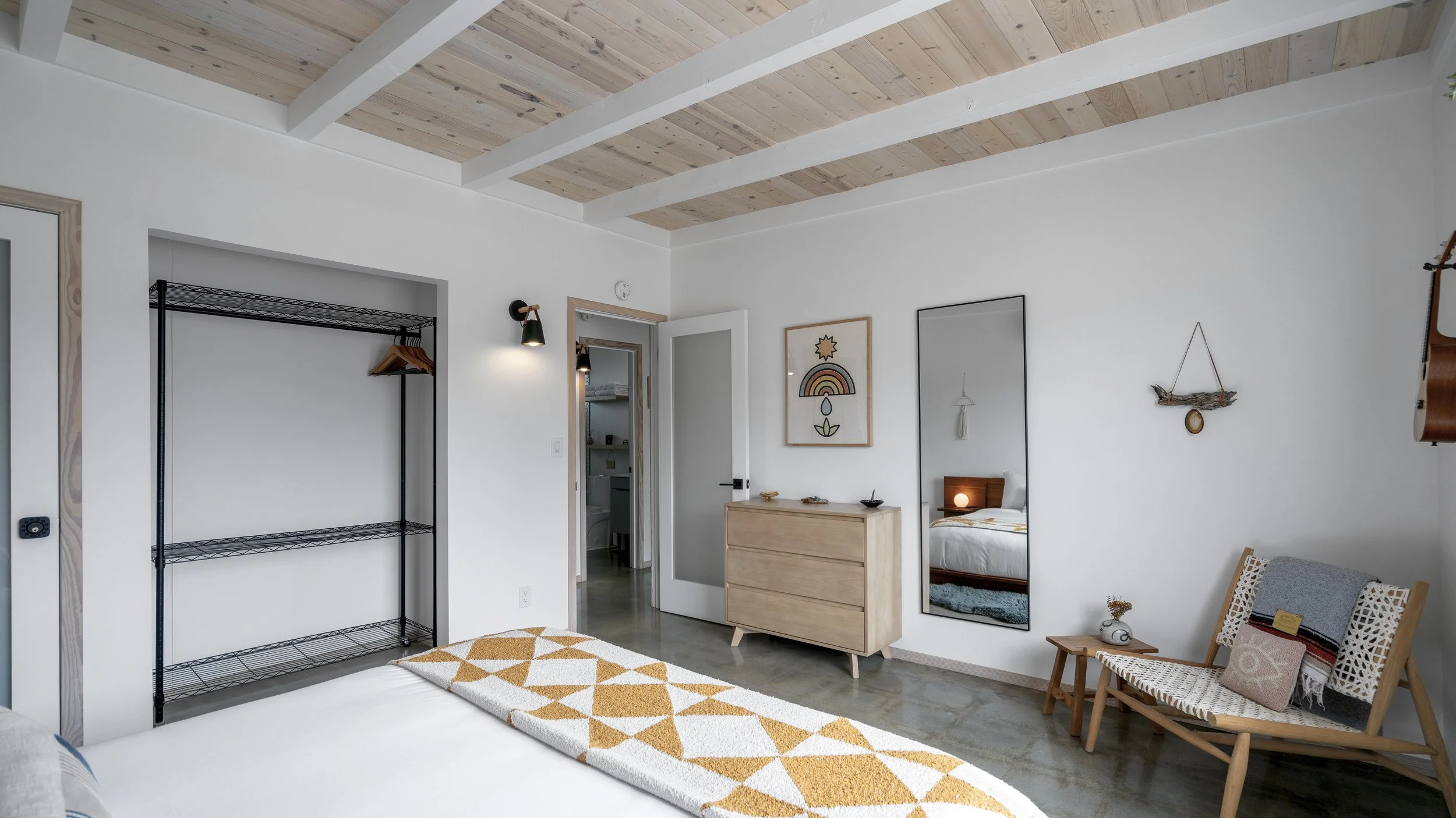 A minimalist bedroom with white walls, wooden ceiling beams, a bed with a mustard and white geometric patterned blanket, a wooden dresser with artwork above it, a full-length mirror, a wooden bench with pillows, and a metal shelving unit in the corne