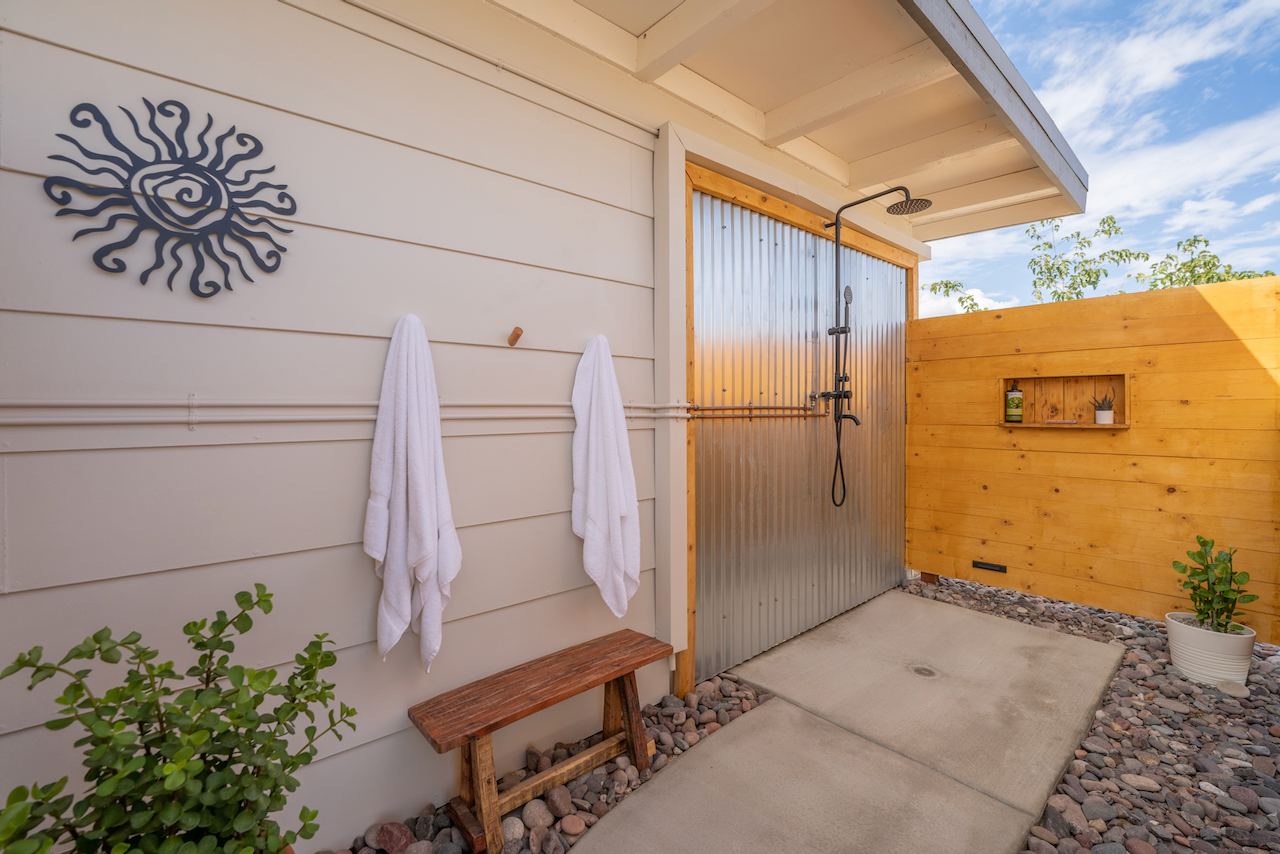 An outdoor shower area with solar-themed wall art, two white towels hanging on hooks, a wooden bench, potted plants, and a wooden privacy wall, under a clear blue sky.