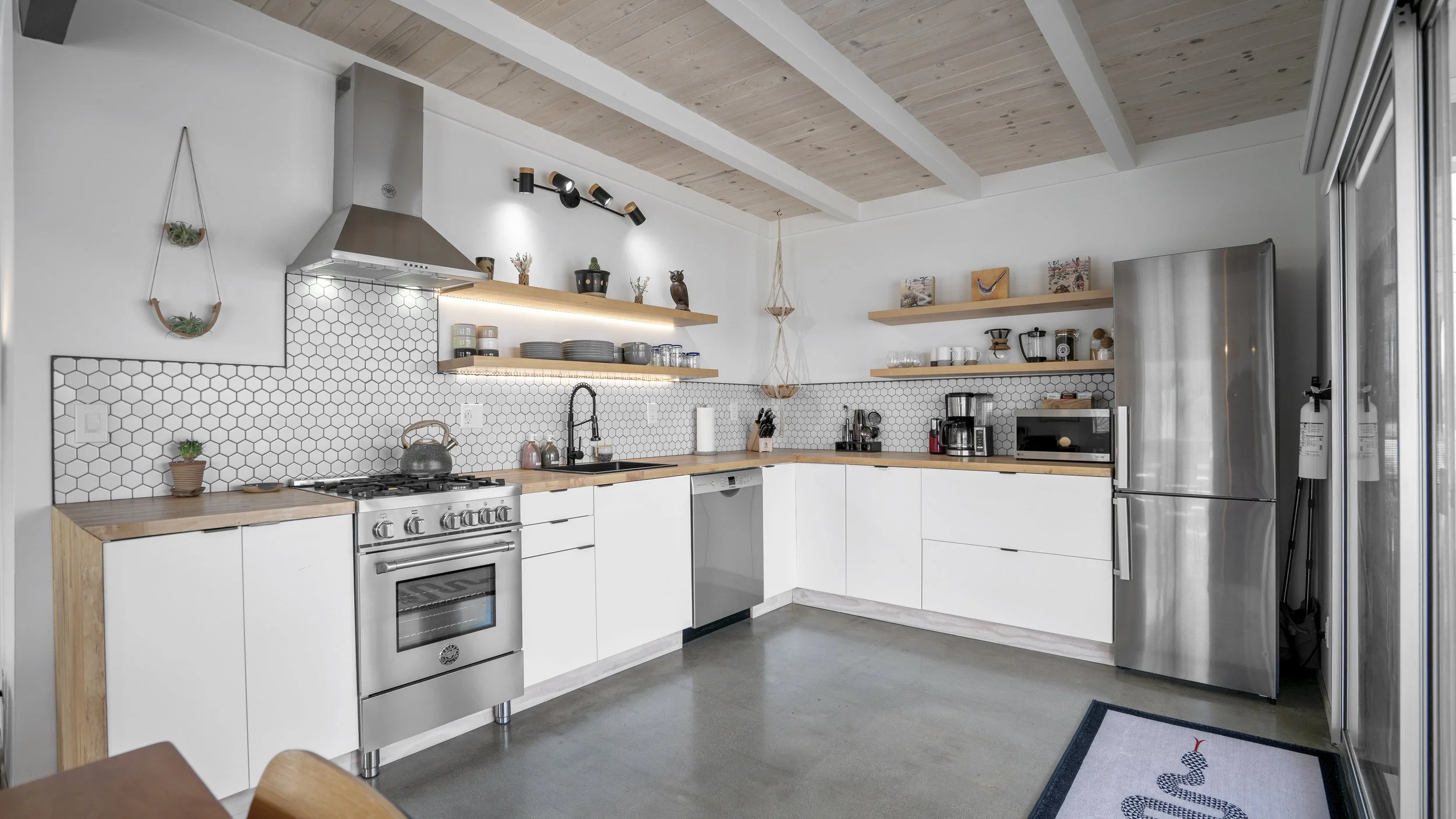 Modern kitchen with white cabinets, wooden countertops, and a stainless steel refrigerator. Open shelves with dishes, cups, and decor. Hexagon tile backsplash and a wood-paneled ceiling.