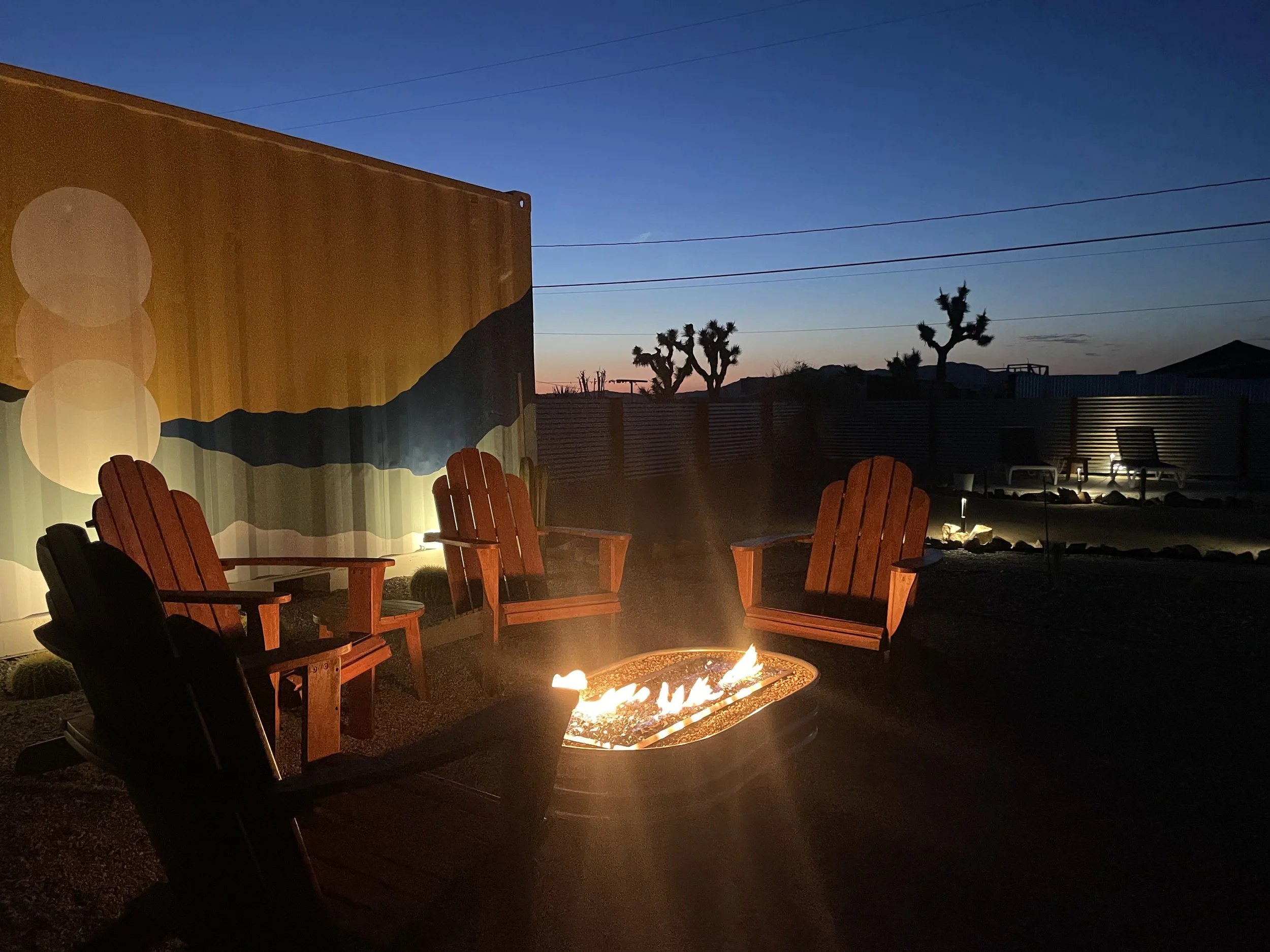 A backyard at dusk with four wooden Adirondack chairs around a lit propane fire pit, desert plants, and Joshua trees in the background, with a painted wall and a fenced perimeter.