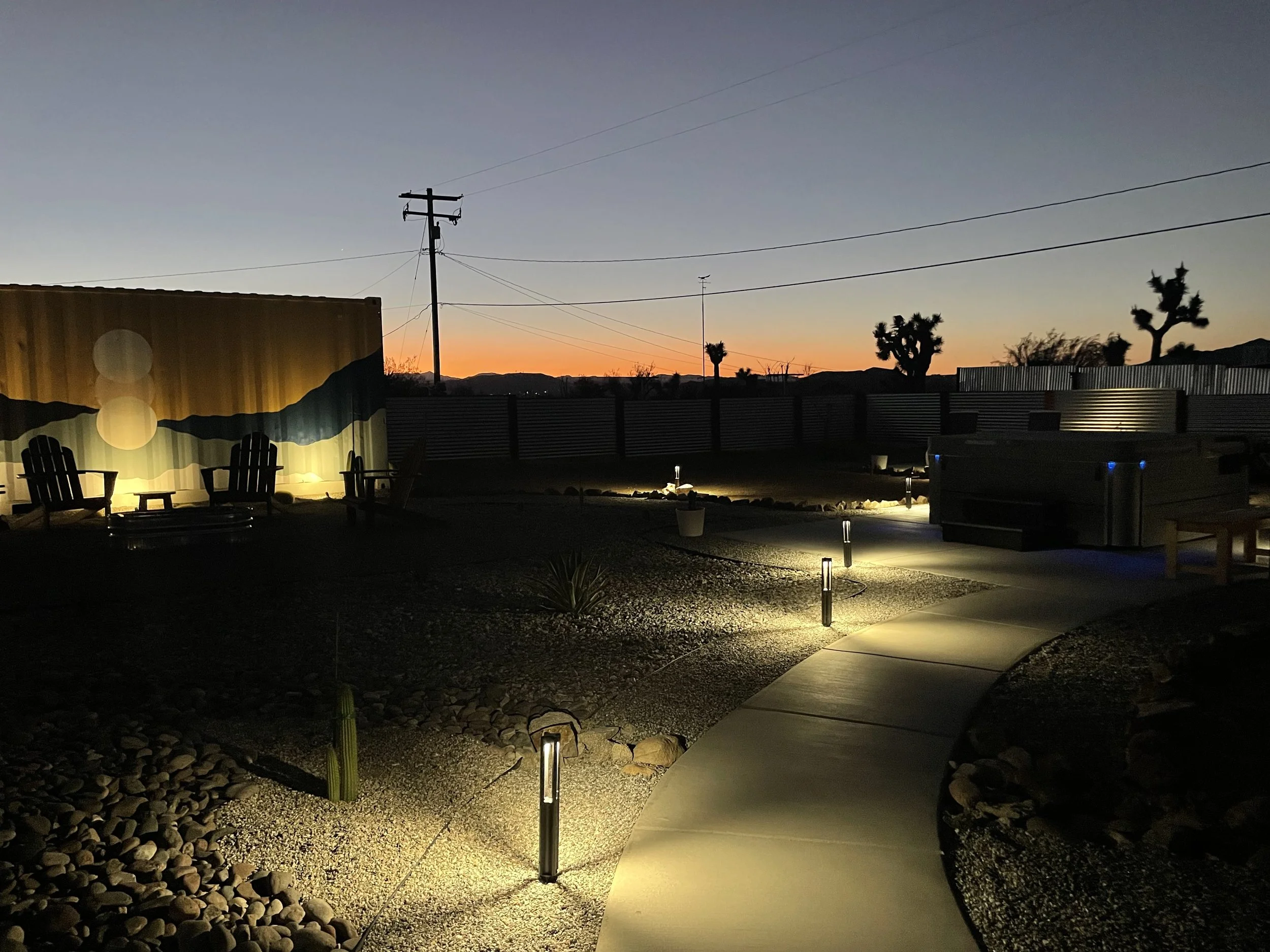 A backyard at dusk with a curved concrete pathway, small outdoor lights illuminating the path, desert plants, a painted wall with Adirondack chairs, and mountains on the horizon against a twilight sky.