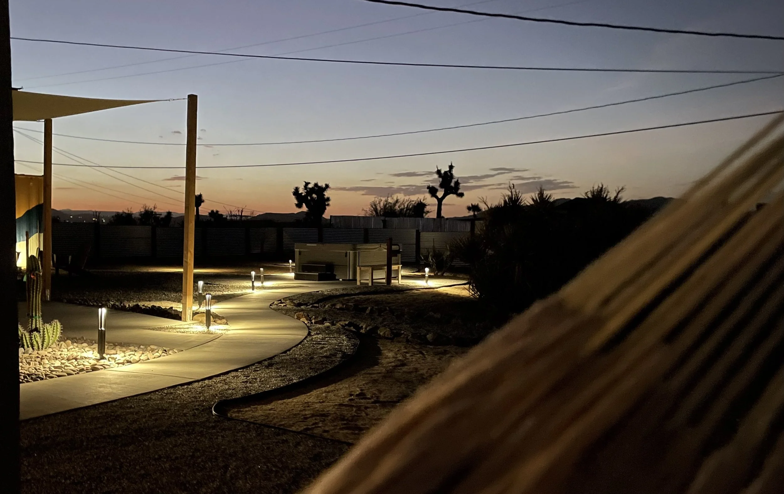 A desert landscape at dusk with a pathway lit by small ground lights, cacti, and Joshua trees in the background, and mountains in the distance.