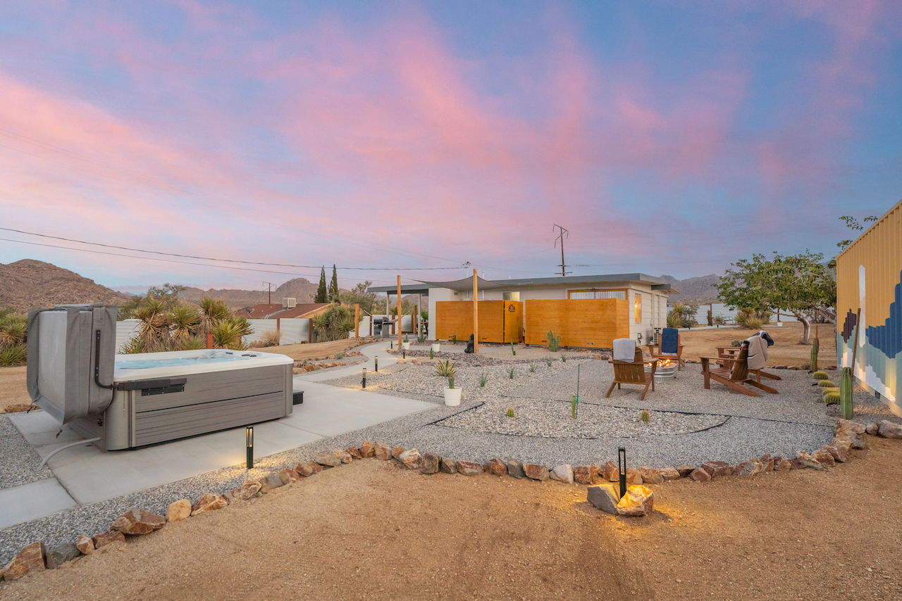 A backyard scene during sunset with an outdoor hot tub, gravel pathways, a fire pit with chairs, and desert landscaping.