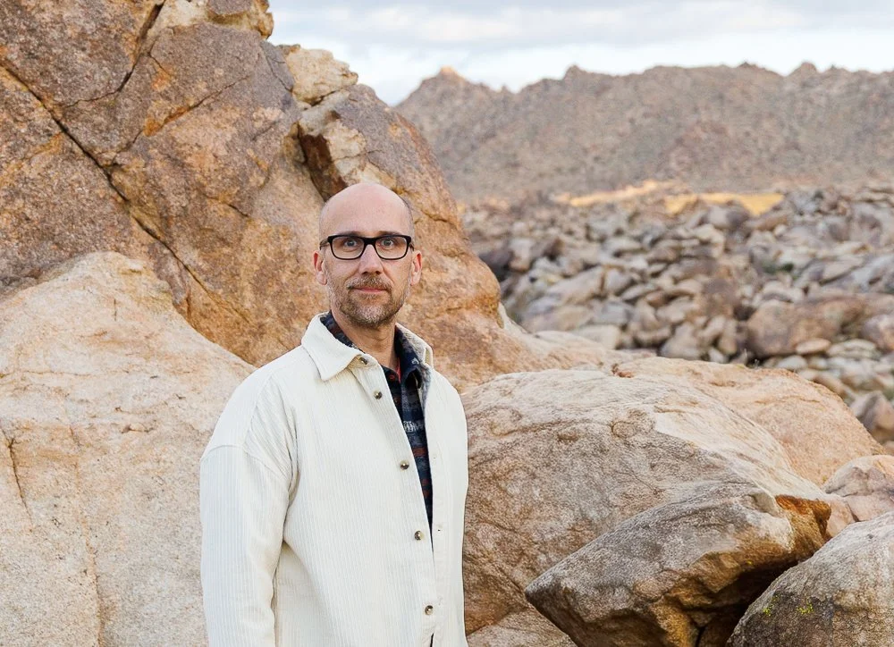 A man with glasses and a beard wearing a white jacket standing in a rocky desert landscape with mountains in the background.