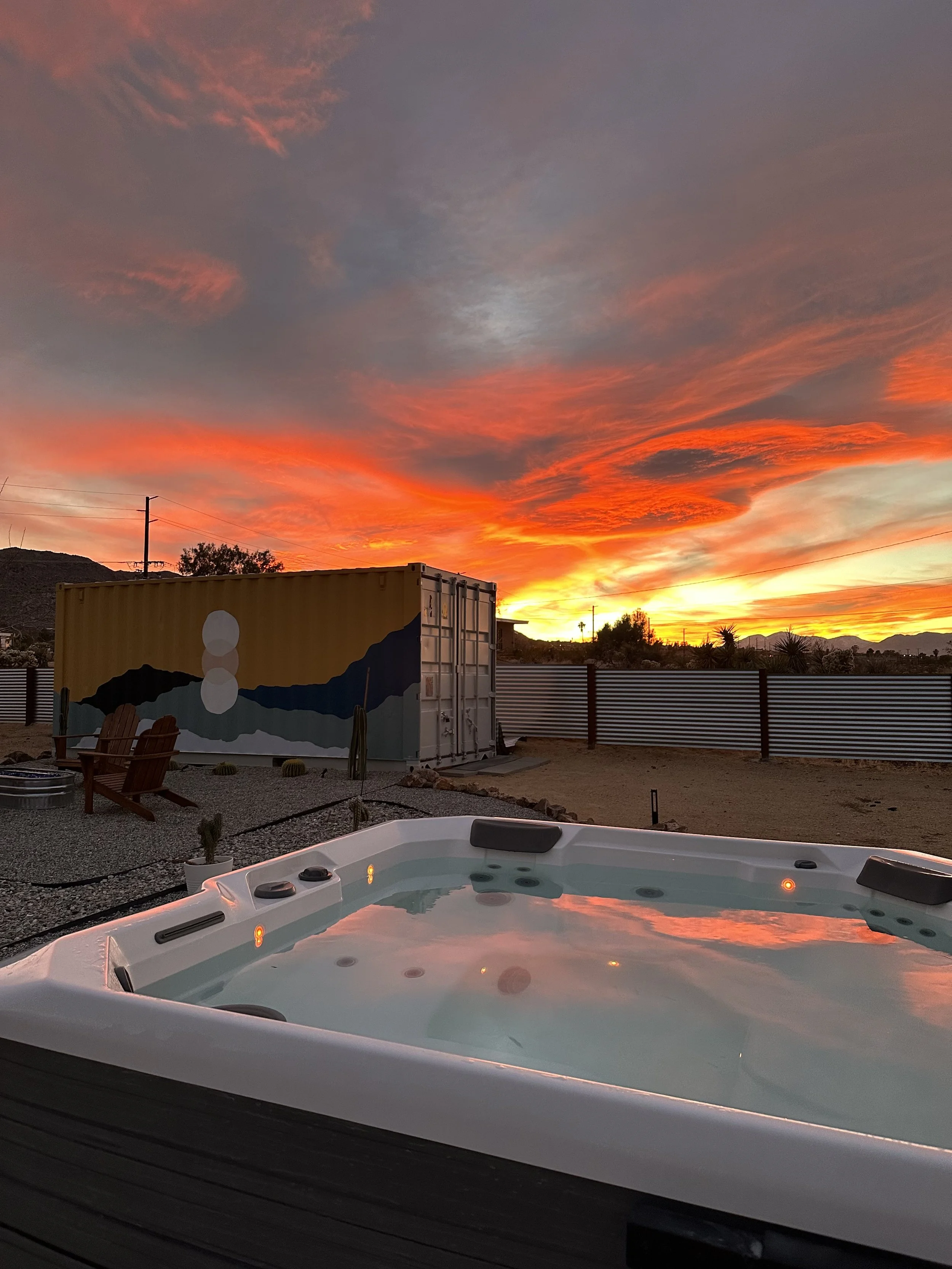 Outdoor hot tub with a sunset sky featuring vibrant orange, yellow, and pink clouds, a painted shipping container, two wooden chairs, cacti, and a metal fence.