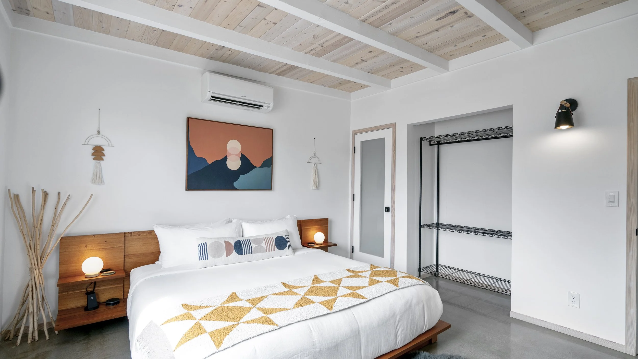 Modern bedroom with minimal decor, white walls, wooden ceiling, bed with white linens and a patterned throw, artwork above bed, small bedside lamps, open closet with wire shelving, and a decorative wall light.