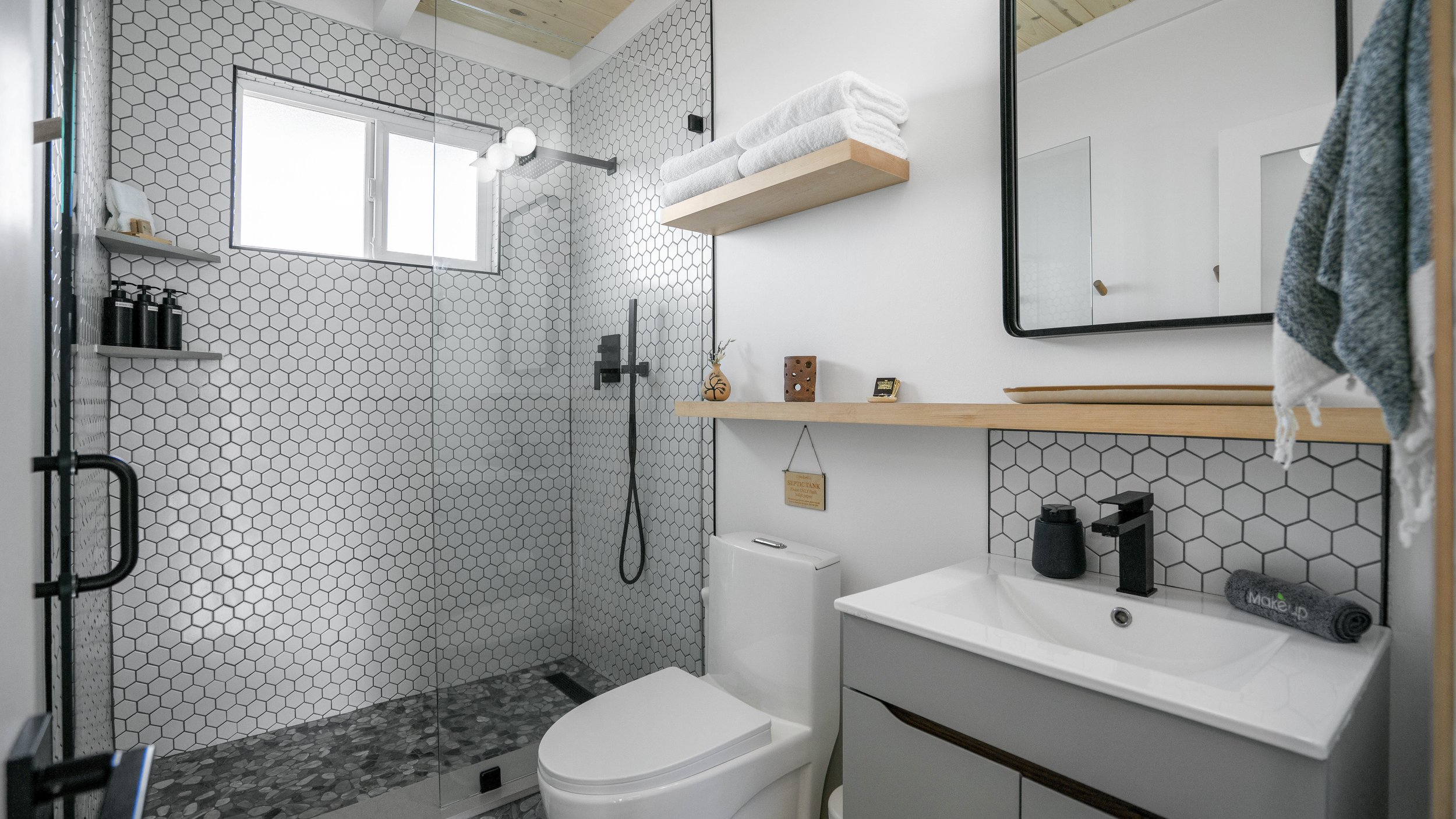 Modern bathroom featuring a walk-in shower with hexagon tiles, a window, a white toilet, a vanity with a rectangular sink, a mirror, and wooden shelves with towels and decorative items.