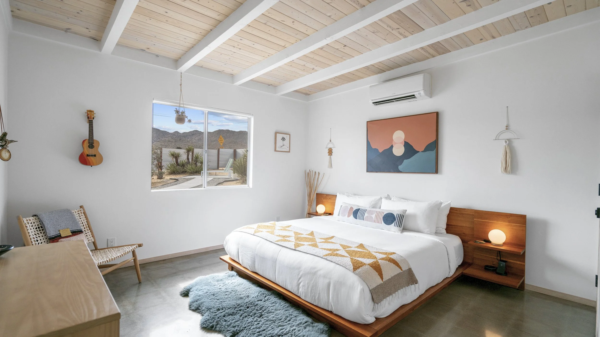 Modern bedroom with white walls, wooden ceiling, and mountain view through window. Features a bed with white bedding, decorative pillows, a wooden headboard and side tables with lamps, a wall art piece, and hanging ornaments. Also includes a guitar o