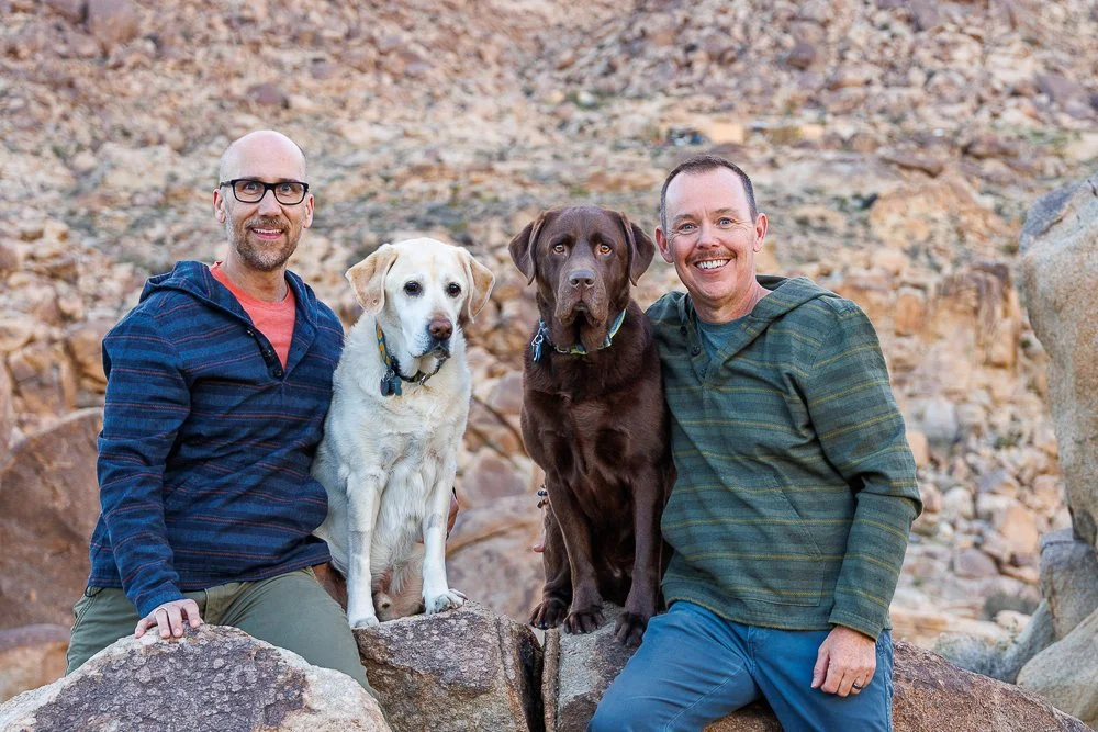 Two men and two dogs outdoors on rocky terrain, one man in glasses and beard, one man smiling, with one light-colored and one dark-colored dog.