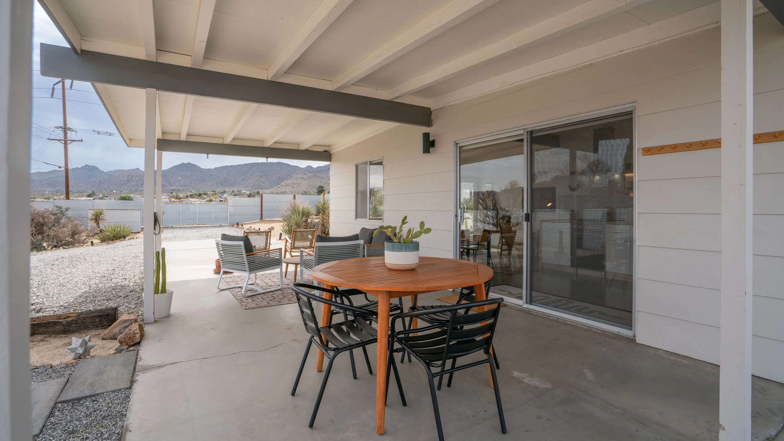 Outdoor patio with seating area including a round wooden table with a cactus plant, black chairs, and additional chairs and sofa in the background, with desert landscape and mountains in the distance.