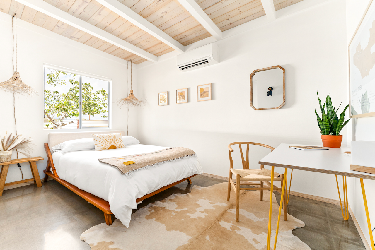 A bright bedroom with white walls, a wooden bed with white bedding and pillows, a window with a view of a tree outside, hanging woven lamps, and a desk with a houseplant and a chair.