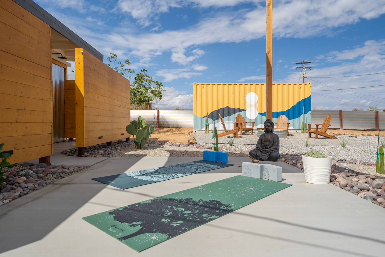 Outdoor yoga space with mats, a Buddha statue, painted container wall, Adirondack chairs, desert plants, and a partly cloudy sky with utility pole