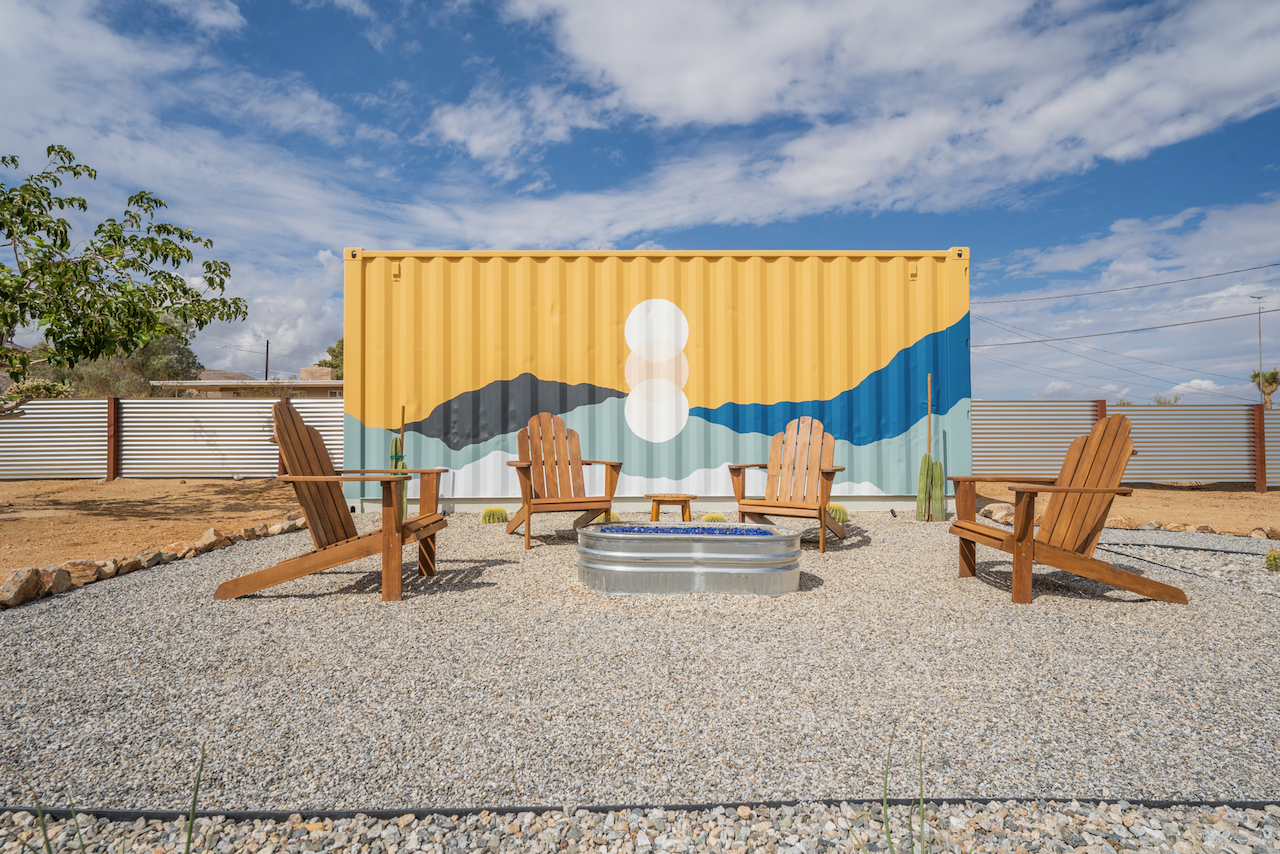 Outdoor seating area with four wooden Adirondack chairs around a small fire pit on gravel, with a colorful mural of mountains and clouds painted on a shipping container in the background under a partly cloudy sky.
