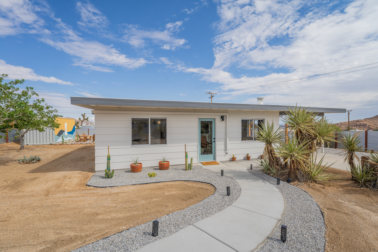 Small white house with a light blue door, two front windows, and desert landscaping with cacti and succulent plants under a partly cloudy sky.