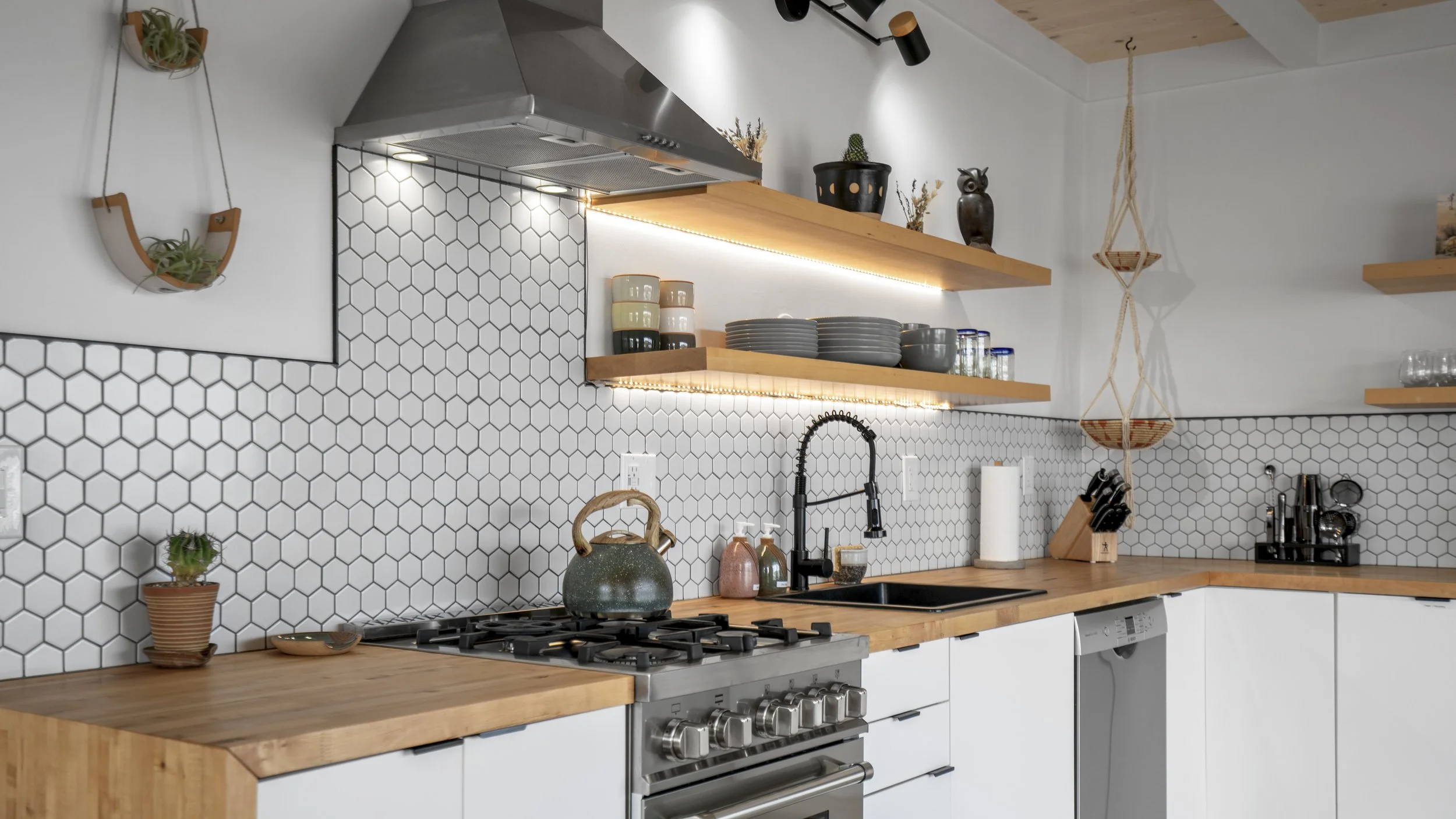 Modern kitchen with white cabinets, wooden countertops, hexagon tile backsplash, open wooden shelves with dishes and decor, stovetop with teapot, sink with black faucet, hanging basket, potted plants, and kitchen utensils.