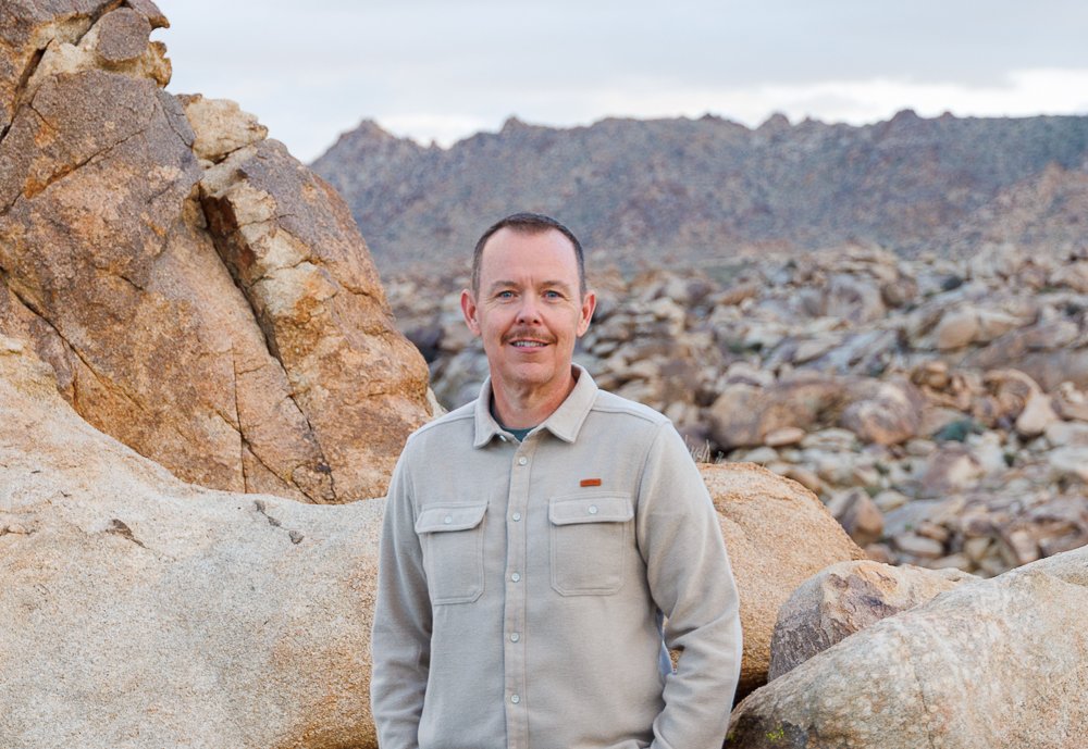 A man standing among large rocks in a desert landscape with mountains in the background