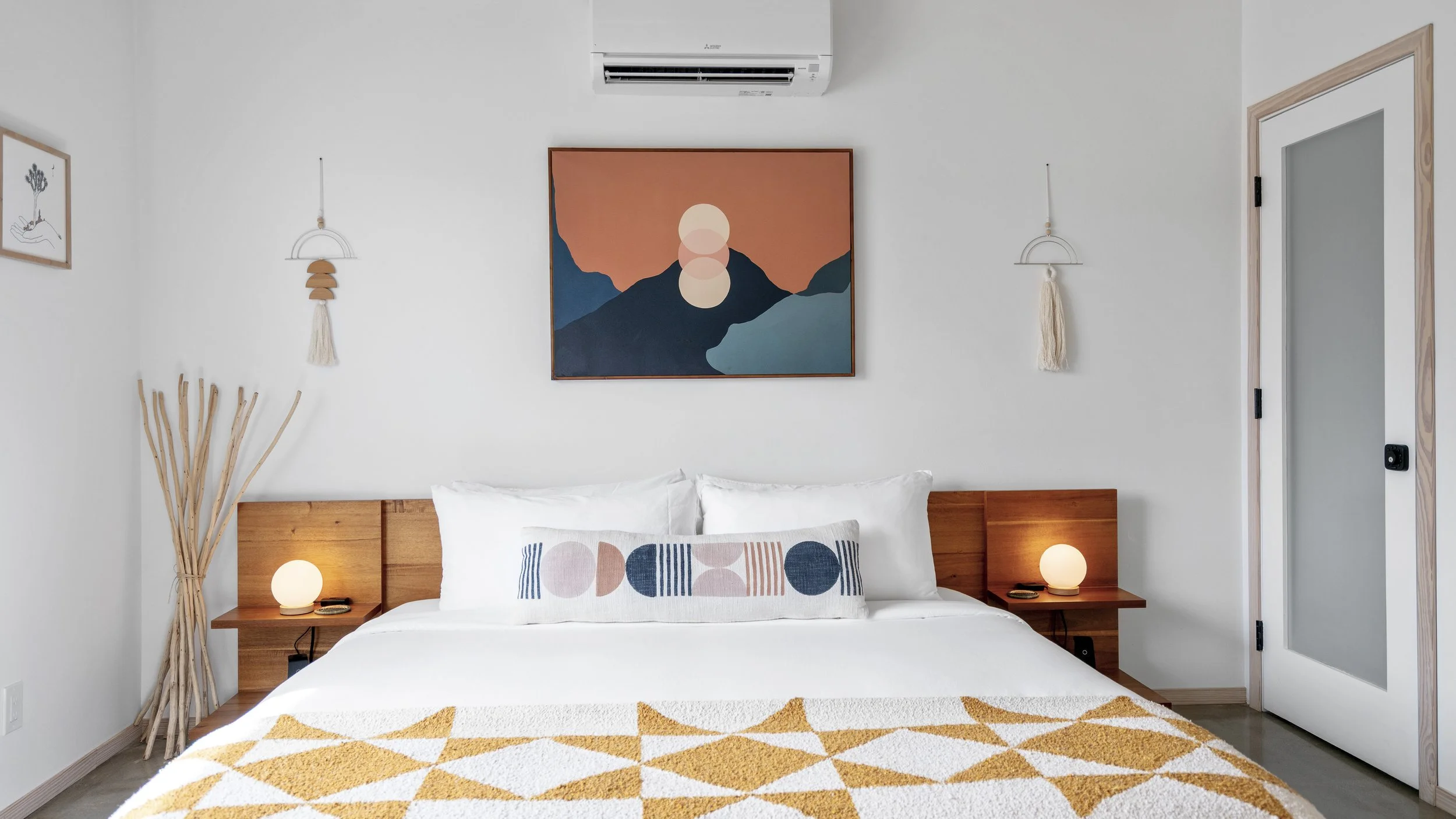 A bedroom with white walls and wooden accents. The bed has white bedding, a decorative pillow with geometric patterns, and a yellow and white geometric patterned blanket. Two small bedside tables with round lamps are on either side of the bed. Artist